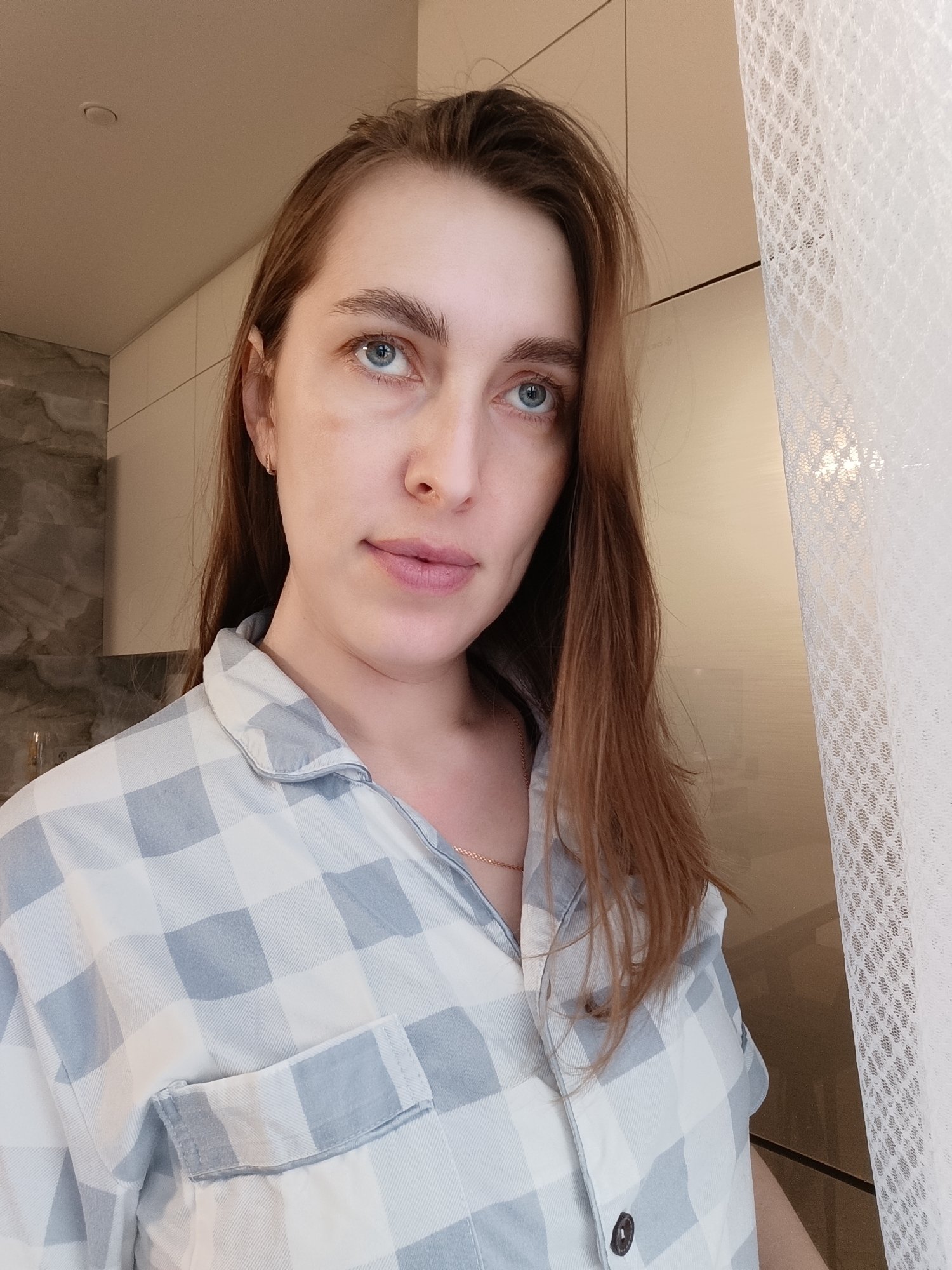A woman with fair skin, light brown hair, and blue eyes is depicted in a close-up shot. She's wearing a light blue and white checkered button-up shirt, possibly pajamas. A small gold earring is visible in her left ear, and a delicate gold chain is around her neck.
The background includes a section of a room with light-colored cabinets and a grey marble-like wall texture in the upper left corner. To the right of the woman, a white lace curtain partially covers what appears to be a window or doorway.
While the image provides details about the subject and immediate surroundings, there is no information present to confirm the location as Chaikovsky, Russia. Therefore, it's not possible to verify the city location based on the given image.
