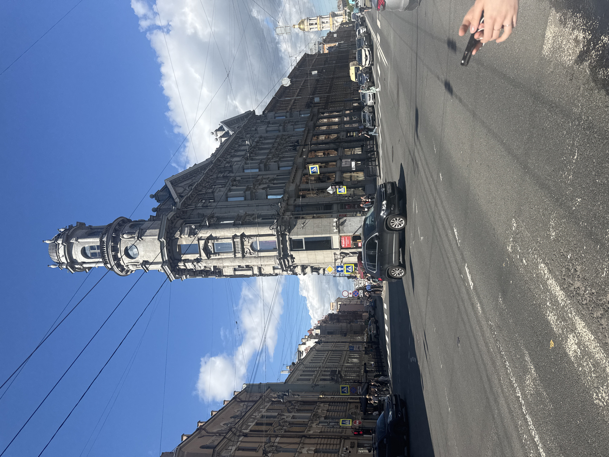 The image depicts a wide city street in Saint Petersburg, Russia, on a bright, partly cloudy day. The focal point is a striking, multi-story building with a distinctive wedge shape and a prominent tower topped with domes. The architecture is ornate, featuring numerous windows, decorative elements, and balconies. The building is situated at a corner intersection, with other buildings lining the street on either side, exhibiting a similar, older European style.
The street is paved with asphalt and marked with white lane lines. Several vehicles are visible, including cars, a van, and a bus, indicating moderate traffic. Pedestrians are present on the sidewalks and crossing the street at designated crosswalks. Traffic signals and street signs are visible. Overhead, a network of power lines crisscrosses the sky. In the distance, a golden-domed building, likely a church or cathedral, is visible, adding to the characteristic Saint Petersburg skyline. The overall scene conveys a sense of urban activity within a historic city setting.