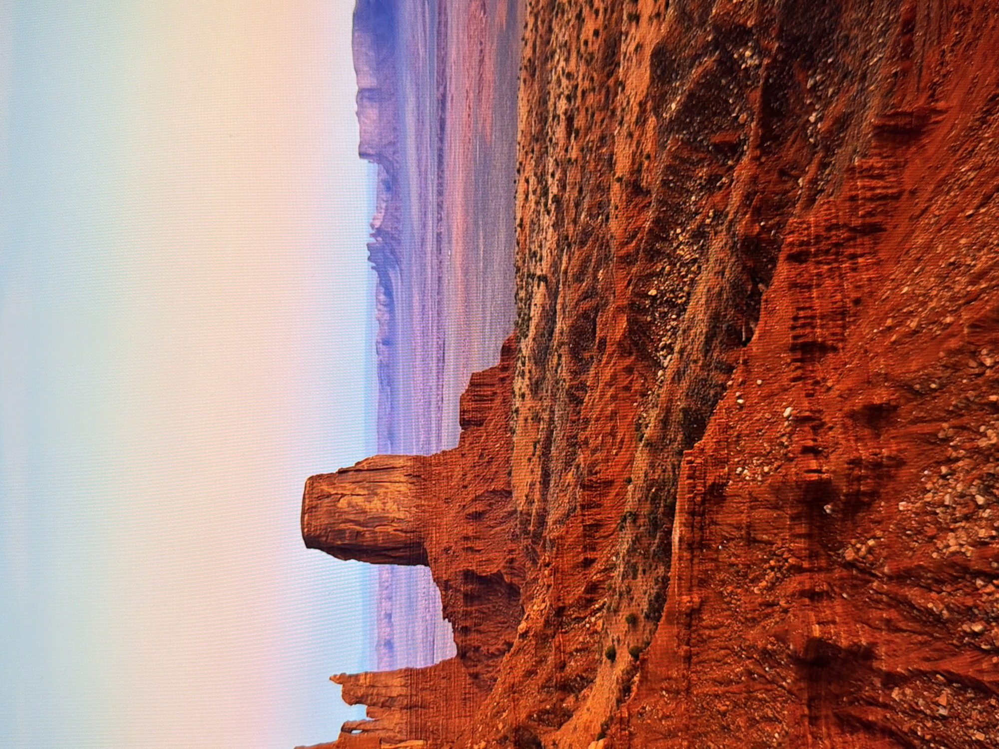 This is a photograph of Monument Valley, located on the Arizona-Utah border in the United States, not in Kyiv, Ukraine. The image depicts a vast desert landscape with prominent sandstone buttes. The sky above is a gradient of pale blue and soft orange, suggesting either sunrise or sunset. In the foreground, the rugged, red-orange terrain is characterized by steep, eroded rock formations and sparse vegetation. The midground features several large, iconic buttes, including one of the most famous, the Mittens. Beyond the buttes, the landscape stretches into a flat, dusty plain, with distant mesas and buttes fading into a hazy, purple-hued horizon. The overall impression is one of grand scale and natural beauty, with a focus on the geological formations and the atmospheric lighting.