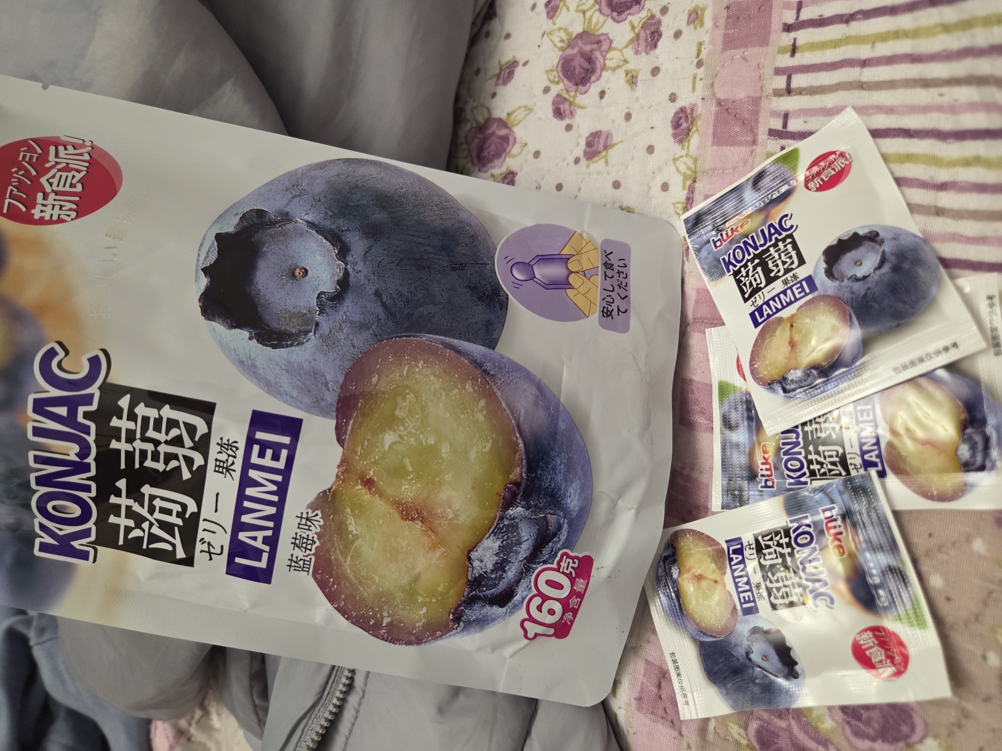 The image depicts a package of "KONJAC LANMEI" blueberry-flavored jelly snacks. A larger, open package lies next to three smaller, individual servings of the same product. The packaging features an image of a sliced blueberry. The setting appears to be a domestic environment, as the snacks are resting on patterned fabric that may be bedding or a tablecloth. There's also a gray, possibly zippered jacket or blanket visible in the lower left corner. There is no indication of the location being Pleven, Bulgaria.