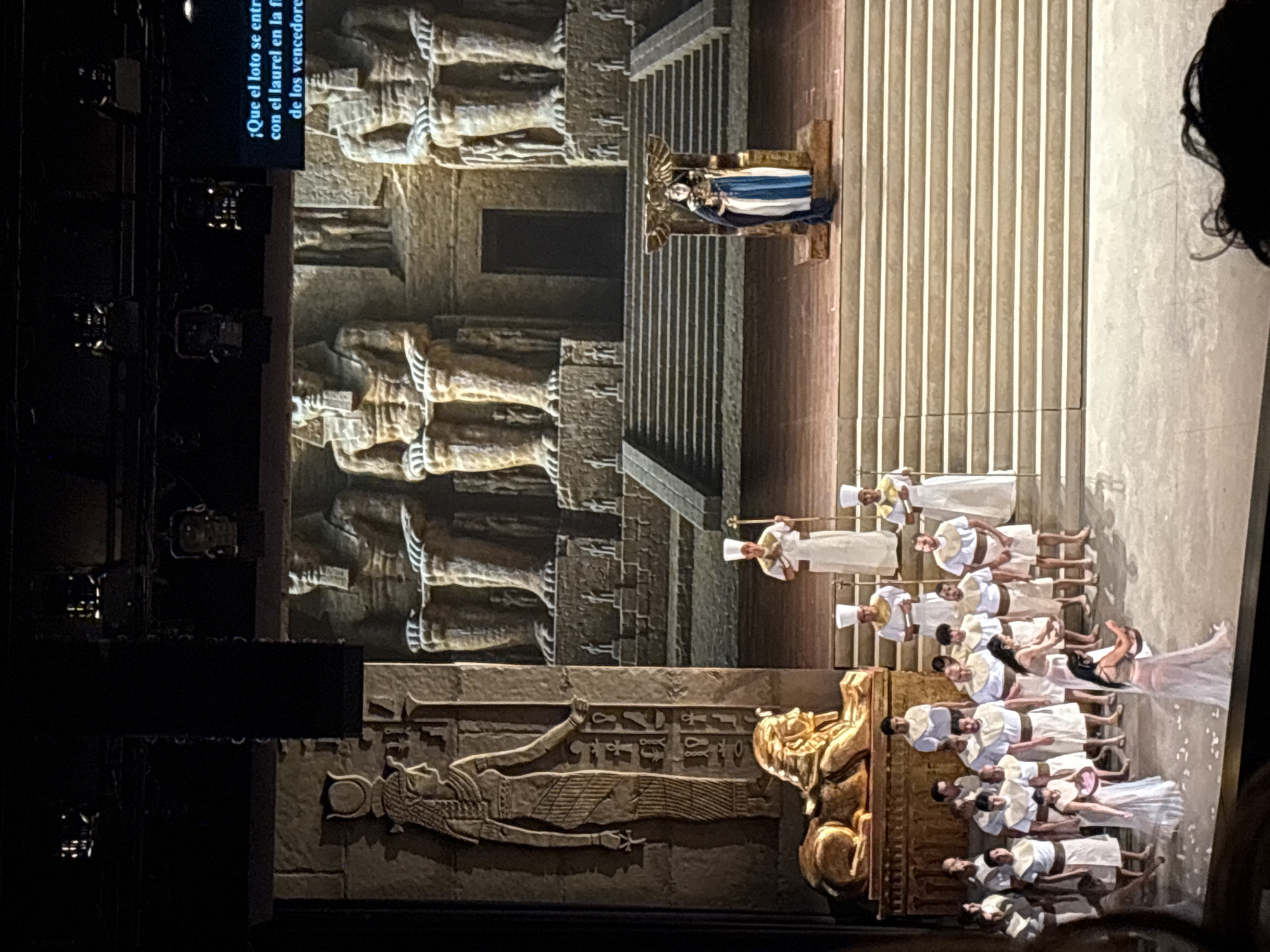 This image depicts a theatrical stage production, possibly an opera, likely taking place indoors.
Setting: The stage is elaborately decorated with elements resembling ancient Egyptian architecture. There are large textured walls adorned with hieroglyph-like carvings and statues. A staircase leads to a raised platform where a figure dressed in blue and white attire sits on a golden throne.
Subjects: A group of performers dressed in white robes and headdresses are positioned near the base of the stage.
Action: The performers appear to be part of a processional or a tableau, possibly singing or chanting. The figure on the throne is the focal point of the scene, potentially a queen or goddess.
People: Predominantly appear to be performers on stage in costumes. Additionally, a person's head is visible on the far right of the frame.
Objects: The stage set includes a throne, architectural elements resembling ancient Egyptian structures, costumes, lighting fixtures, and potentially props.
Environment: The lighting appears to be dramatic, with spotlights highlighting the performers and architectural details.
Location: This stage production took place in San Borja, Peru.