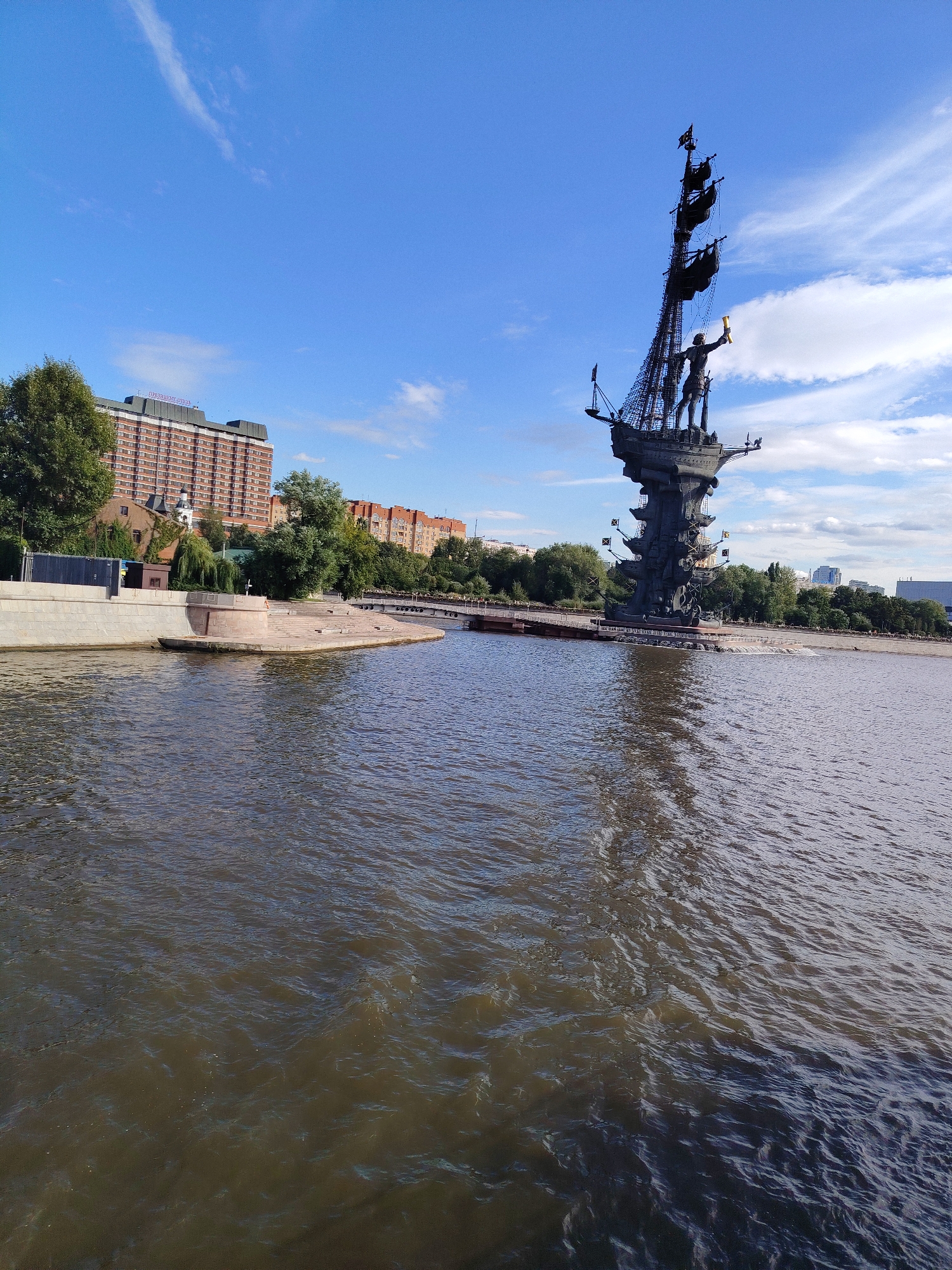A vertical, outdoor, medium shot captures a bronze statue of Peter the Great in Moscow, Russia, on a sunny day. The statue, which resembles a ship with a man standing on top holding a scroll, is situated on the Moskva River. The river is in the foreground, its water a murky brown with ripples. Behind the statue, lush green trees line the riverbank. A large, brick building with many windows and a rooftop sign is visible on the left side of the frame. The sky above is a clear blue with a few wispy white clouds.