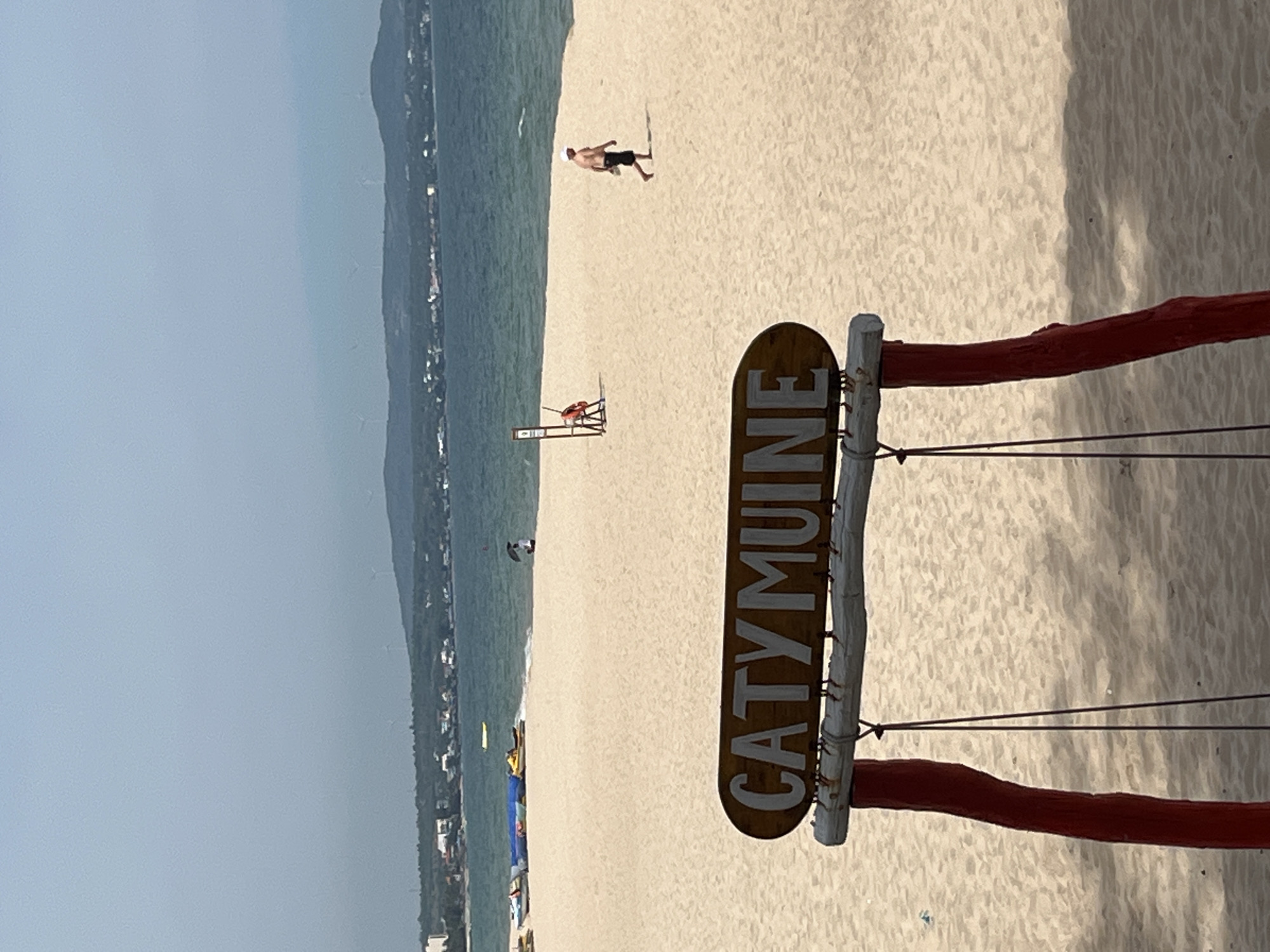 The image depicts a sunny day at a sandy beach in Mũi Né, Vietnam. In the foreground, a weathered wooden sign with the words "CATY MUINE" painted in white letters is prominently displayed, supported by rustic wooden posts and red beams. The sign is partially obscured by a thin wire.
The beach itself is wide and covered in light-colored sand. In the middle ground, a lone person, a man, walks across the sand, wearing dark shorts and a white cap. Further down the beach, a red lifebuoy is mounted on a wooden stand. The ocean, a tranquil shade of blue-green, stretches out towards the horizon. Along the coastline, a strip of green vegetation and scattered buildings suggest a coastal town. The overall atmosphere is relaxed and typical of a beach vacation destination. The time of day appears to be late morning or early afternoon, given the brightness of the sun and the distinct shadows cast on the sand. The weather is clear and pleasant, ideal for a beach outing.
