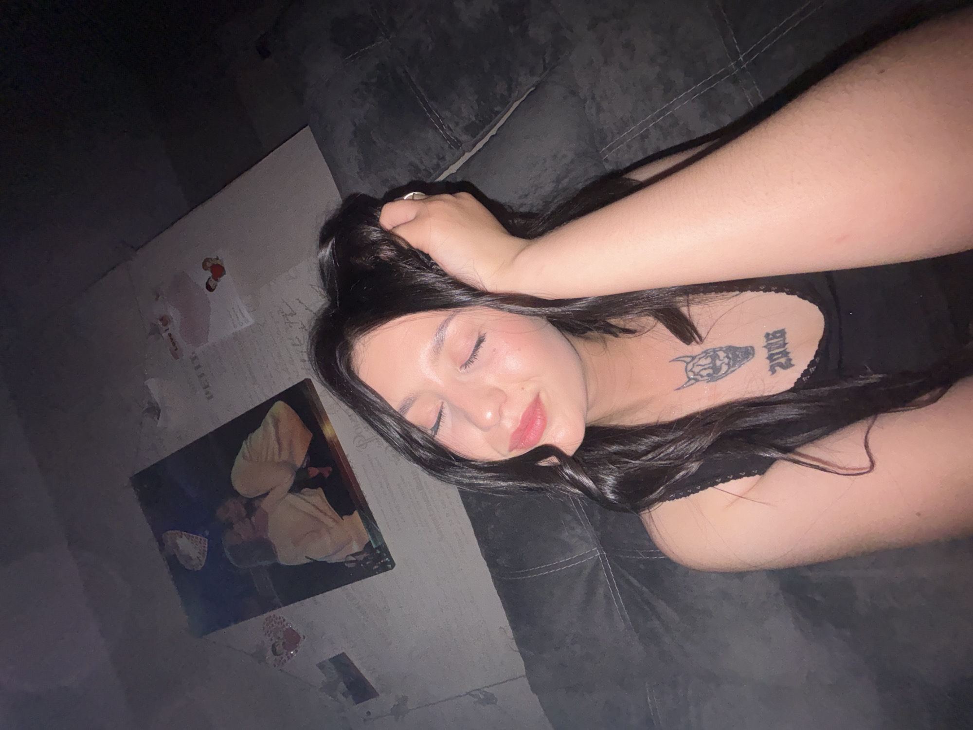 The image shows a young woman with long, dark hair, her eyes closed, and her hand resting on her head. She is wearing a black top with lace trim and has a tattoo of a horned skull and the numbers "2006" on her chest. The background appears to be a dark, possibly upholstered surface, like a couch. On the wall behind her, there are several framed photographs and decorative heart-shaped cutouts. One of the photographs appears to be a close-up of two people embracing. The overall lighting is dim, suggesting an indoor, possibly nighttime setting. There is no information available in the image to determine the location in the city of Chaikovsky, Russia.