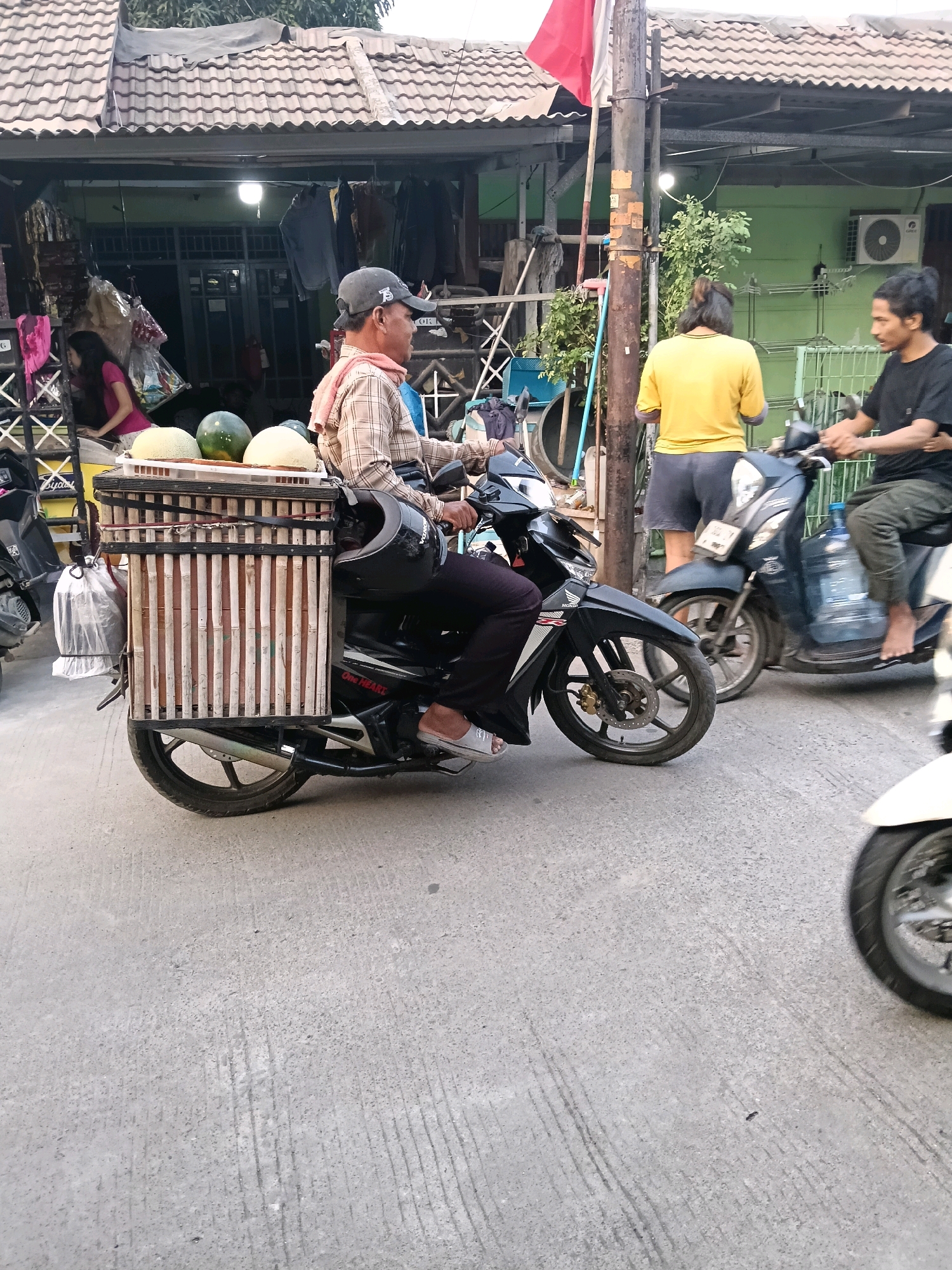 In Jatimulya, Indonesia, a man wearing a checkered shirt, a baseball cap, and dark pants rides a black scooter. The scooter is equipped with a large, bamboo-constructed carrier filled with what appear to be melons. The man is carrying a pink cloth draped around his neck. Behind him, another scooter is visible with a person in a yellow shirt and dark shorts, and further in the background, a person in a black t-shirt and dark pants is also on a scooter. The scene takes place on a street with buildings lining the side, featuring corrugated roofs and open storefronts displaying merchandise. The ground is paved with concrete.