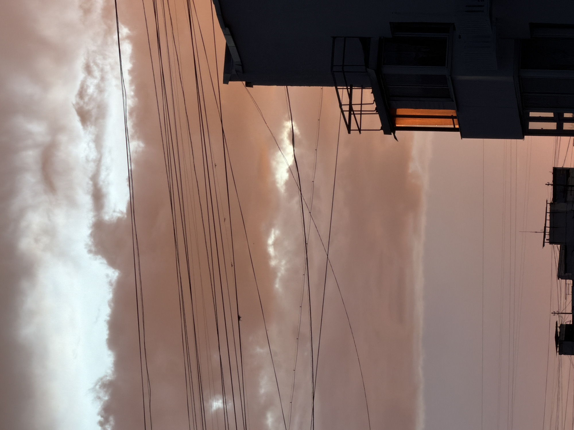 The image shows a view of the sky with clouds and power lines, with the silhouettes of buildings against the colorful sunset sky. The clouds are dramatic and varied, with bright white areas contrasting with softer, pinkish-orange tones. The sky suggests it is either sunrise or sunset, with warm light illuminating the clouds. Several power lines crisscross the frame, creating a geometric pattern that divides the sky.
In the foreground and extending into the background are the dark, angular shapes of buildings. One building on the right has balconies and windows, with one window reflecting the warm sunset light. Another building appears to be under construction or in a state of disrepair, with exposed scaffolding and incomplete structures. The overall scene evokes a sense of urban landscape at dusk or dawn, with a blend of natural beauty in the sky and man-made structures.
The location is identified as Khimki, Russia. There is no visible text in the image. The image's orientation is vertical, with the horizon line running horizontally across the frame. The mood is somewhat dramatic and contemplative, due to the stark contrast between the dark buildings and the vibrant, illuminated sky.