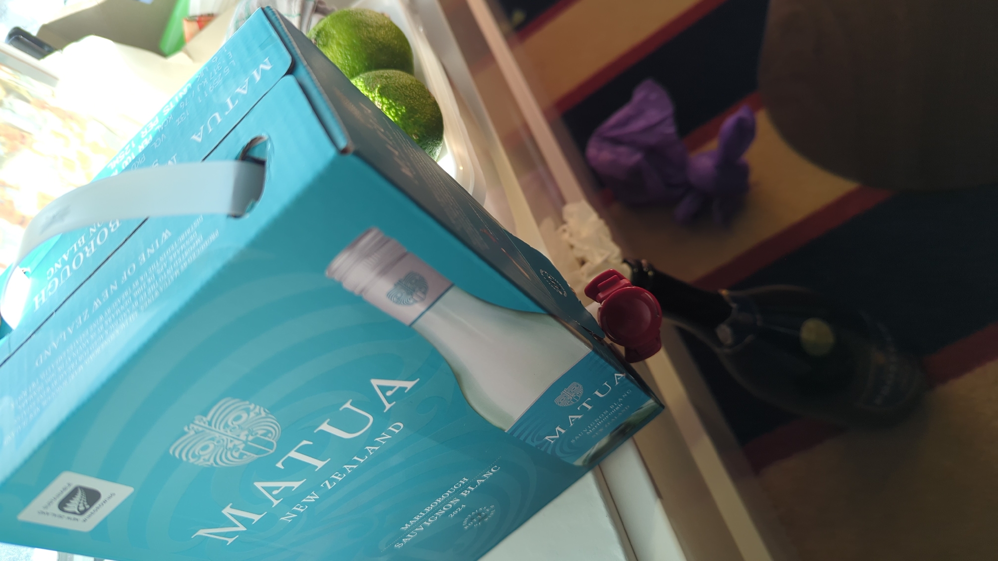 The image depicts a cluttered surface, possibly a table or shelf, with several items present. Dominating the scene is a light blue cardboard box for a wine brand called "Matua," originating from New Zealand. The box is tilted and features an illustration of a wine bottle and brand logos. To the right of the box, two green limes are visible on a white plate. In the foreground, a dark glass bottle with a red stopper is positioned vertically, its label mostly obscured. A purple fabric or object lies behind the bottle. The background is blurred, but suggests an indoor setting with hints of a rug with red and yellow stripes on the floor. The scene is illuminated by natural light, casting shadows and highlights. The location is identified as Larkollen, Norway.