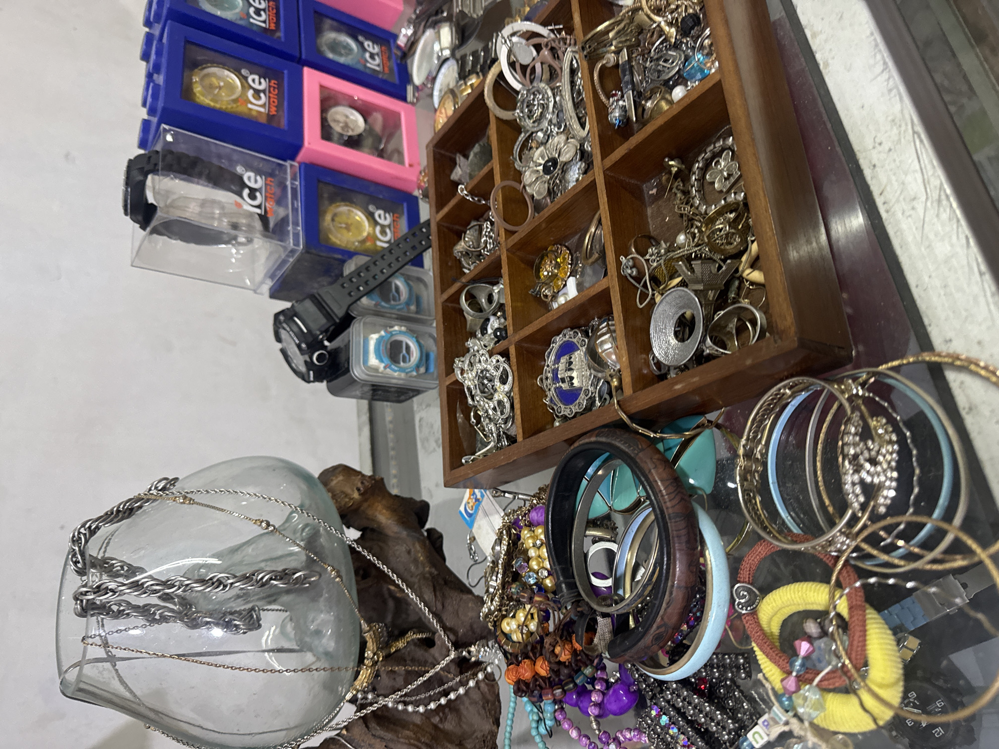 A flat-lay photograph captures a collection of jewelry and watches displayed on a glass surface. In the upper left quadrant, a large glass vase, appearing to be part of a repurposed artistic piece, holds several thick, silver-toned chains. A thin, gold-toned chain is draped over the edge of the vase. To the right of the vase, stacked watch boxes with the "Ice Watch" logo are visible, some containing watches. Below the vase and extending towards the right, a dark brown wooden compartmentalized tray is filled with various pieces of jewelry. These include rings, necklaces, pendants, bracelets, and brooches, with a notable blue and silver enamel pendant and several silver rings in the upper left compartment. Scattered across the glass surface in front of the wooden tray are more bracelets, some appearing to be bangles, and other assorted jewelry items. The lighting suggests an indoor setting. The scene does not contain any people or indicate any specific activity or interaction beyond the static display of items. The origin of the photograph is attributed to Denpasar, Indonesia.