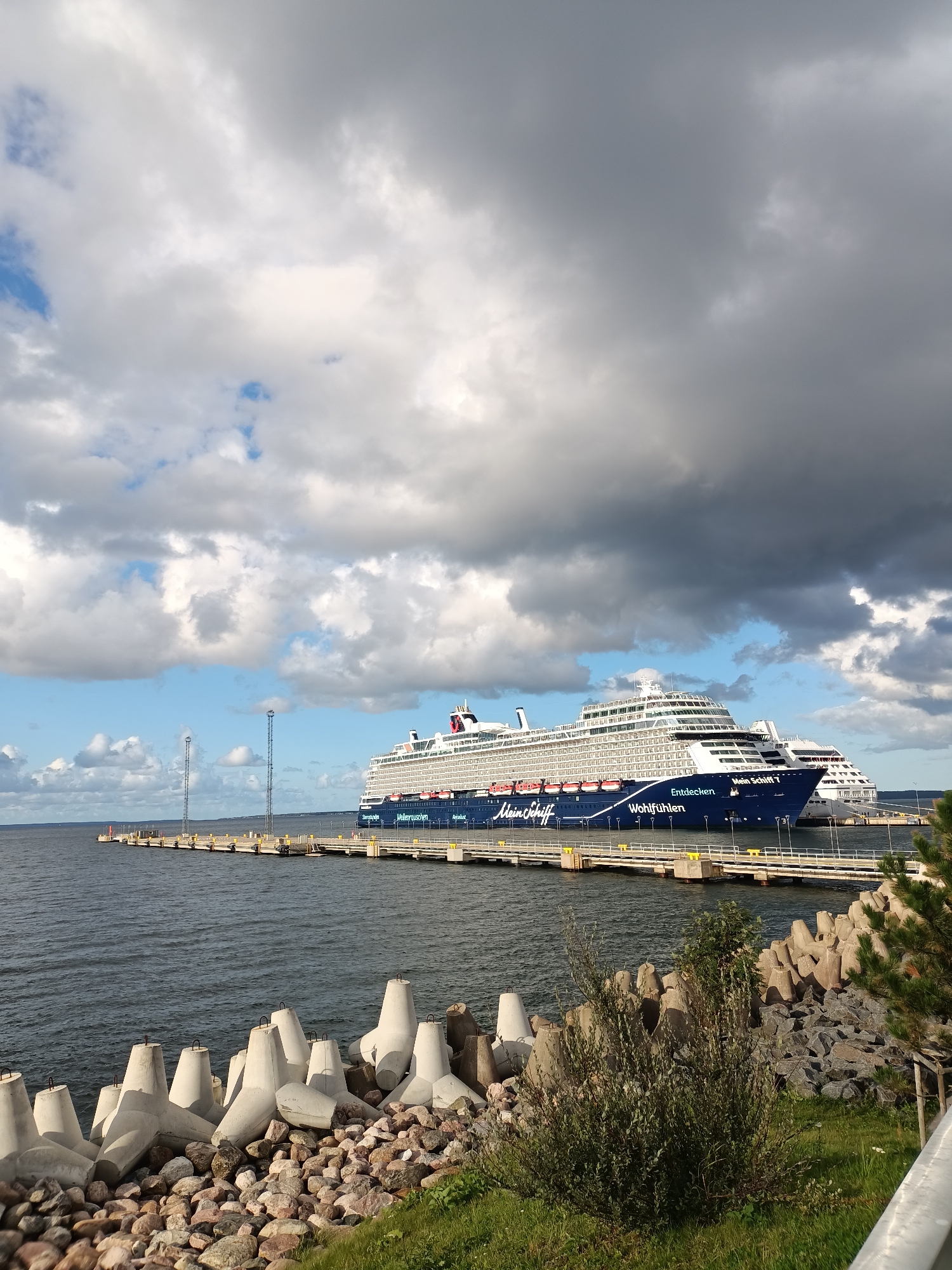 In Tallinn, Estonia, on a cloudy day, a large blue and white cruise ship named "Mein Schiff 1" is docked at a pier. The ship is a prominent feature in the image, with its name clearly visible on the side. The pier extends into the dark, rippling water, and is lined with yellow safety railings and tall poles with lights. In the foreground, a rocky shore is adorned with an array of concrete tetrapods, some of which are partially submerged in the water. A smaller, white cruise ship is visible behind "Mein Schiff 1". The sky is filled with dramatic, grey clouds, with patches of blue sky peeking through. The overall scene depicts a typical day at a port.