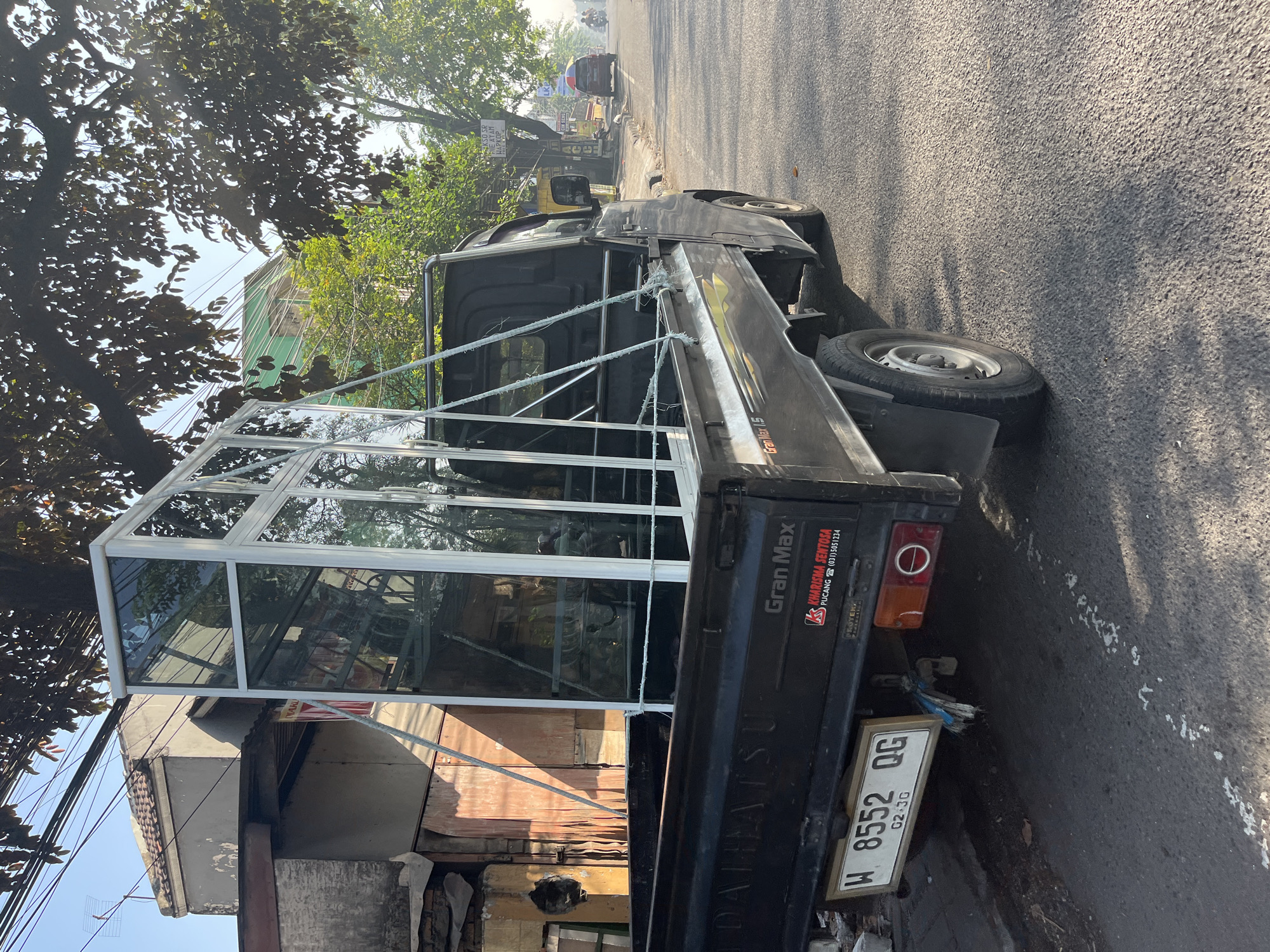 A black Daihatsu Gran Max pickup truck is parked on the side of a street in Candi, Indonesia. The truck bed contains a large, multi-paneled glass structure, possibly a greenhouse or display case, secured with ropes. The truck's license plate reads "W 8552 QG". The street is paved and appears to be a commercial or residential area, with buildings and trees lining the roadside. The sky is bright, suggesting daytime.