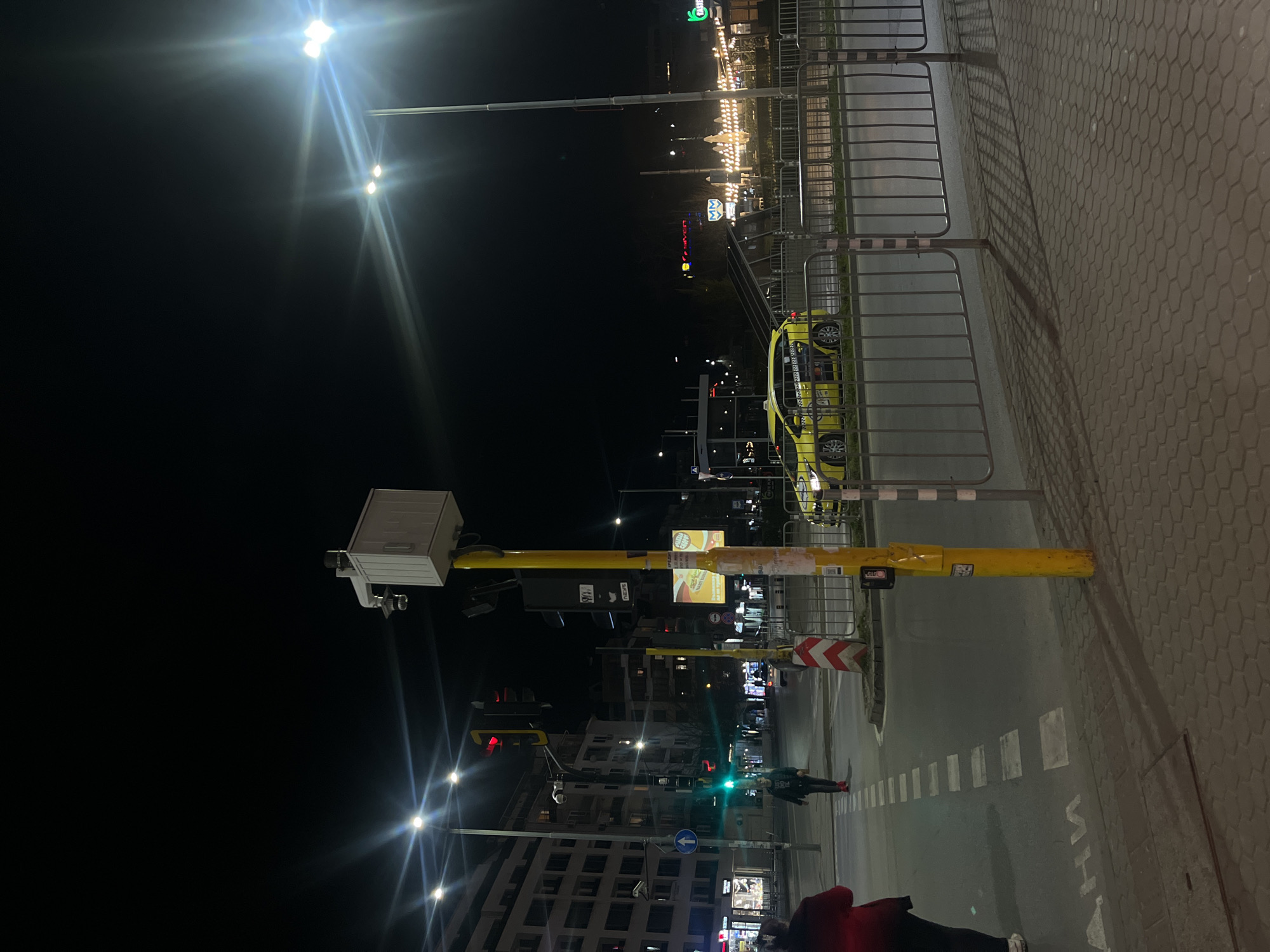 The image depicts a street scene at night in Sofia, Bulgaria. The perspective is slightly rotated counter-clockwise. In the foreground, there's a pedestrian crossing with a yellow traffic signal post. The signal is mounted with a small white box and camera. Further down the street, traffic signals display green and red lights. Pedestrians are visible crossing the street and walking on the sidewalk. In the background, buildings line the street, illuminated by streetlights and shop signs. A yellow taxi is parked along the side of the street next to a metal pedestrian barrier. The overall lighting is dark, with bright spots from streetlights and building illumination.