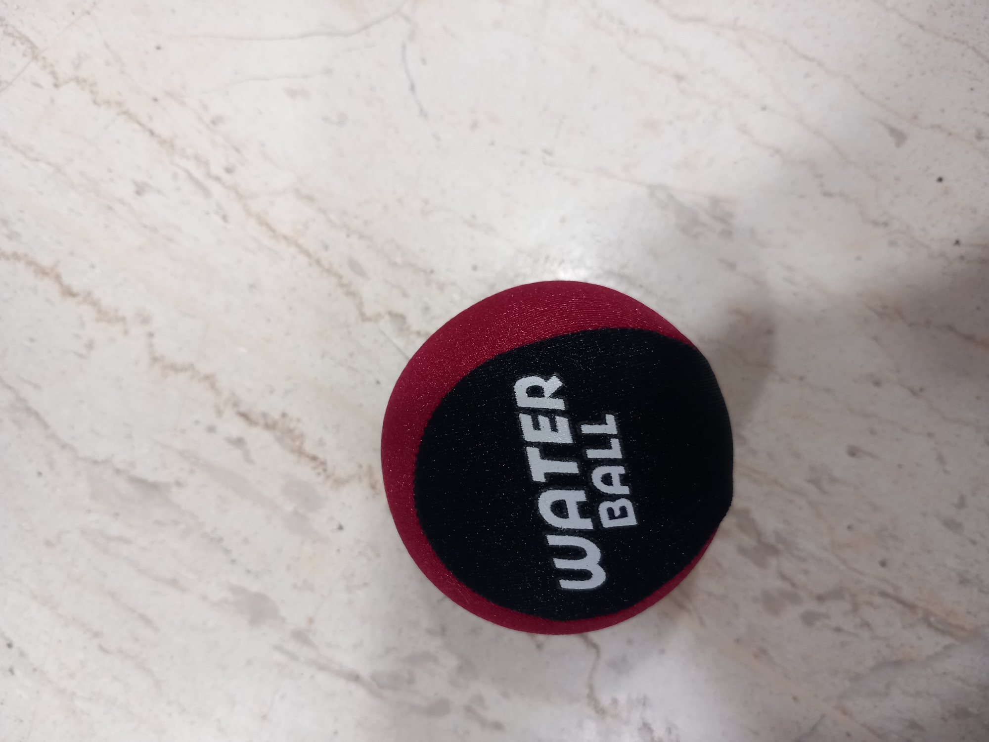 A red and black spherical object, identified by the text "WATER BALL" printed in white on its black surface, rests on a speckled, light-colored surface that appears to be marble or granite. The ball is positioned in the lower right portion of the image, angled slightly such that its entirety is visible. The lighting suggests an indoor setting, with no distinct shadows indicating a specific time of day or weather conditions. The background is a patterned, neutral surface, providing no additional clues about the location. The image is a close-up shot of the object, with the surface taking up most of the frame. The text on the ball is clear and legible, indicating its purpose.