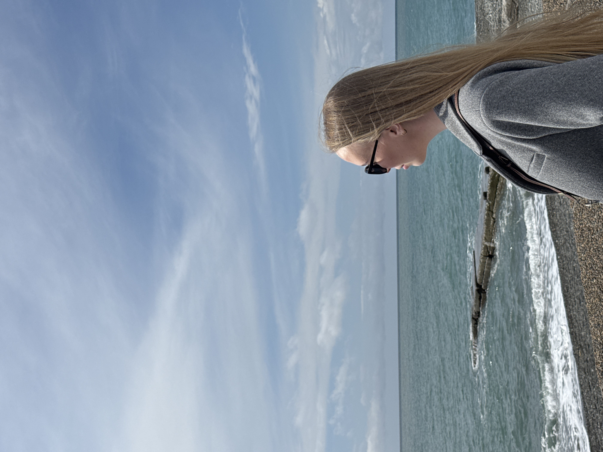 A woman with long blonde hair and sunglasses is standing on a pebble beach, looking down at the sea. She is wearing a gray coat and has a bag slung over her shoulder. The sea is visible in the background, along with a partly cloudy sky. The location is possibly Sochi, Russia.
