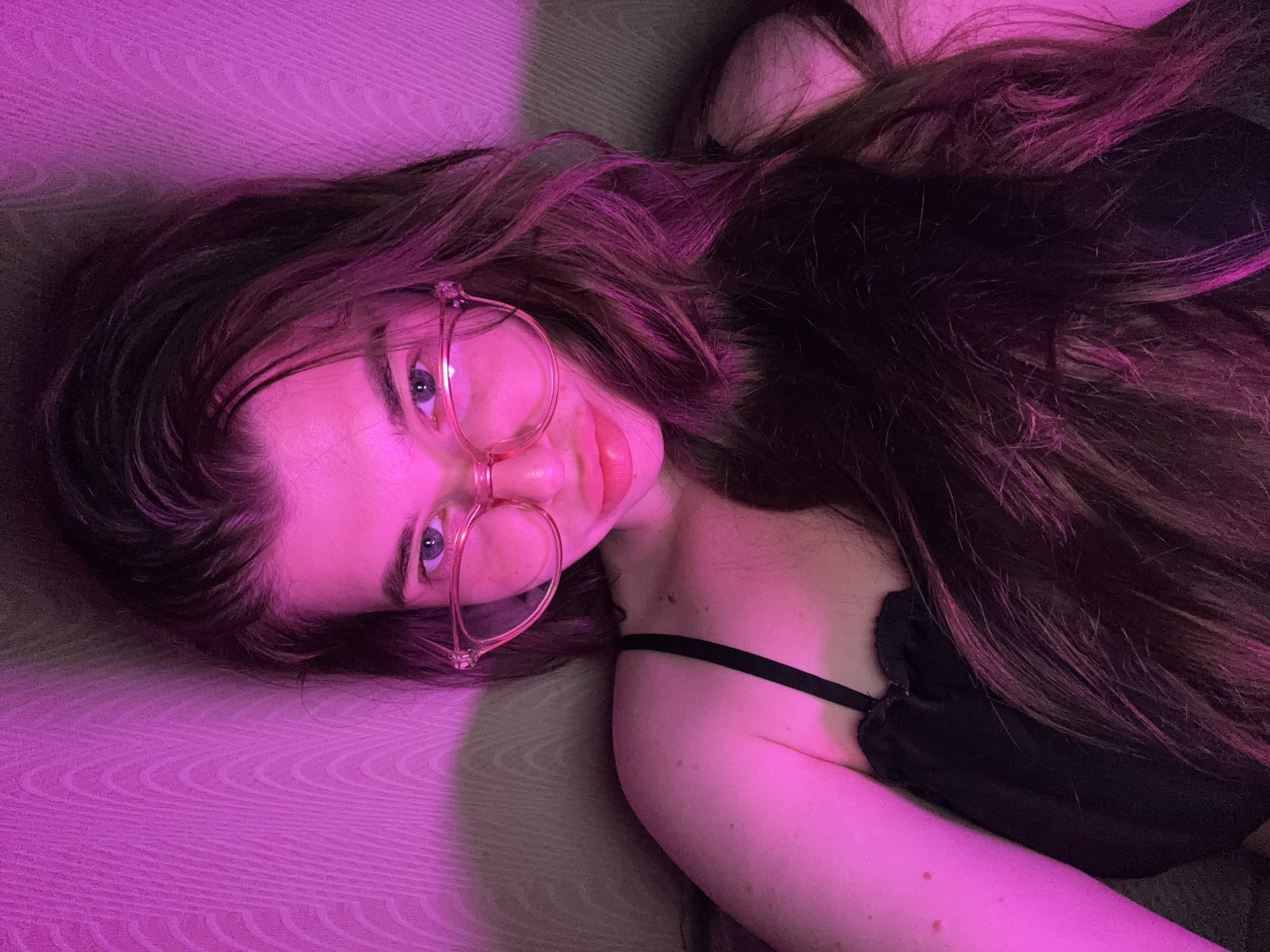 A woman wearing pink-rimmed glasses and a black top is pictured in a dimly lit room with pink ambient lighting. Her long, dark hair cascades around her shoulders. The background appears to be a textured wall, possibly wallpaper, with a subtle wave pattern. The lighting casts a pink hue over the entire scene, including the woman's skin and hair, and creates soft shadows on the wall. The image is a close-up shot, focusing on the woman's face, upper body, and hair. There are no specific details in the image that would allow for a precise location within the city of Мещёры, Russia.