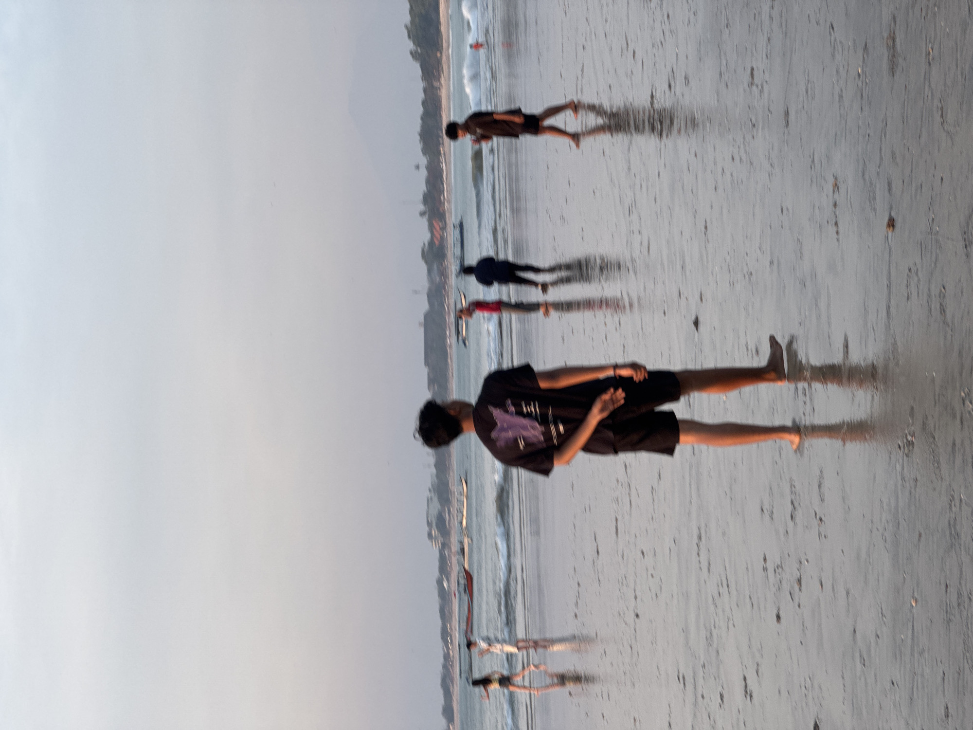 In Kuta Selatan, Indonesia, a serene beach scene unfolds at dusk. The soft light of the setting sun casts a gentle glow on the wet sand, reflecting the figures of people scattered across the shore. In the foreground, a young man stands with his back to the camera, hands clasped behind him, his dark t-shirt and shorts contrasting with the pale sand.
Further back, the gentle waves of the ocean lap at the shore. Several people are visible near the water's edge, some walking and others interacting. A group of boats, likely fishing vessels, are moored in the shallow water, adding to the local ambiance. The background features a hazy coastline with distant hills, suggesting a wider expanse of land beyond the immediate beach. The overall atmosphere is peaceful and tranquil, capturing a typical evening on the Indonesian coast.
