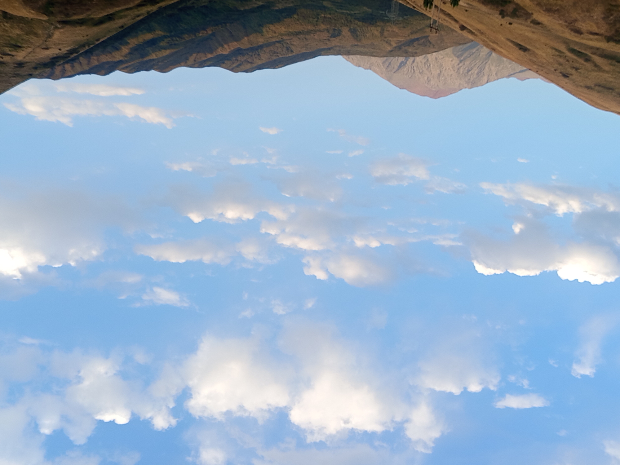 A scenic view of the mountainous landscape near Jardy-Su, Kyrgyzstan, is presented. The upper half of the image is dominated by a clear blue sky adorned with scattered white cumulus clouds, some of which are illuminated by sunlight, creating a bright and airy atmosphere. The lower half of the image features a series of rolling hills and mountains. The closest hills are covered in dry, golden-brown grass, indicative of a semi-arid or late-season environment. Further in the background, the mountains are more rugged and show darker green vegetation in the shaded valleys, with lighter, rocky slopes visible on the sunlit sides. One prominent peak in the mid-ground exhibits a reddish-brown hue on its upper slopes. A utility pole with wires is visible on the right side of the frame, suggesting the presence of infrastructure in the vicinity. The overall impression is one of natural beauty and a vast, open landscape under a dynamic sky.