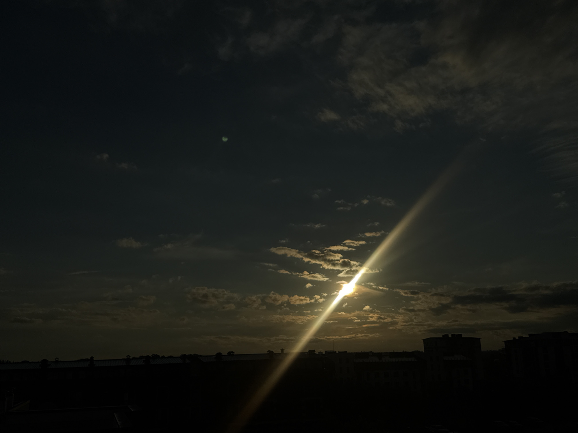 The image depicts a dramatic sky at dusk or dawn over an urban landscape. The sun, a bright point of light with a strong lens flare, is partially obscured by scattered clouds, casting a powerful beam of light diagonally across the frame from the lower left to the upper right. The clouds are varied in texture and density, with some appearing wispy and others thicker, catching the sunlight in shades of yellow and white. The lower portion of the image is dominated by a dark silhouette of a city skyline, featuring the outlines of buildings and rooftops. The environment appears to be an outdoor setting, possibly an elevated viewpoint, with the city stretching out towards the horizon. The overall mood is atmospheric and somewhat moody due to the low light conditions and the striking contrast between the bright sun and the darkened cityscape.