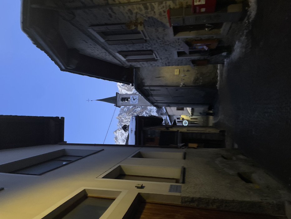 The image, when properly oriented, captures a narrow, atmospheric alleyway in the alpine town of Courmayeur, Italy, during the "blue hour" – either dawn or dusk. Buildings of varying heights and materials, including stone and light-colored plaster, line both sides of the passage, creating a corridor effect.
The focal point in the distance is a distinct church tower with a steeple, topped by a cross, and featuring a clock face. Behind the church, majestic, snow-capped mountains rise under a clear, deep blue sky, indicating an alpine setting. The ground of the alley appears to be paved and shows some patches of snow or ice, suggesting cold weather conditions.
Artificial lighting is visible further down the alley on the right side, casting a subtle glow on the building facades and the ground. One such light is a circular, glowing sign affixed to a building, though any specific text or logo on it is not legible. No people are visible, lending a quiet and tranquil mood to the scene. The architecture and mountain backdrop collectively establish the setting as a historic European mountain village.