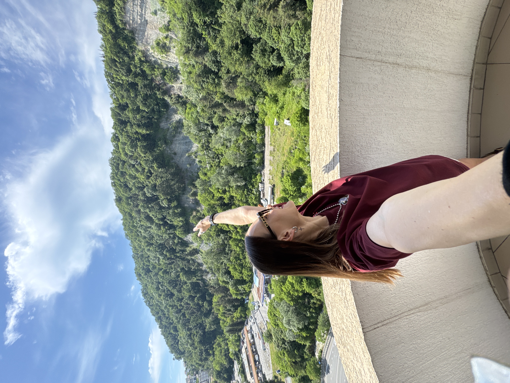 In Sochi, Russia, a woman stands on a balcony, her arm extended upwards as she points towards a verdant mountainside. The mountain is covered in lush green trees, with visible rock formations and layers. Below the mountain, a sprawling urban landscape unfolds, featuring roads, buildings, and what appear to be gas stations. The sky above is a vibrant blue, dotted with fluffy white clouds. The woman is wearing a maroon t-shirt, sunglasses, and jewelry. The perspective of the photo suggests it is a selfie taken from the balcony.