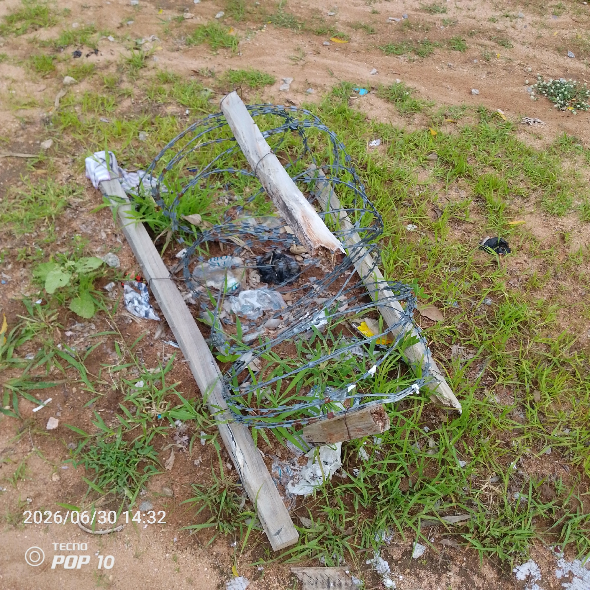 The image depicts a close-up, ground-level view of a collection of discarded materials in an outdoor setting. The primary subjects are two weathered wooden posts, one longer and lying horizontally, and the other shorter, standing upright and angled. Wrapped around these posts is a coil of barbed wire, suggesting it might have been part of a fence or barrier.
Scattered among the posts and barbed wire is a collection of trash, including plastic bottles, crumpled plastic bags, and what appears to be a black plastic bag. Some white, torn fabric is also visible, along with other unidentifiable debris. The ground is a mixture of sandy soil and patches of green grass, indicating an undeveloped or neglected outdoor area.
The scene appears to be set during the daytime, with no strong shadows suggesting a specific time other than it is not dusk or dawn. The weather is not discernible from the image. The presence of trash and the haphazard arrangement of the materials suggest this is a place where waste has been dumped, possibly in an unmanaged area.
Visible text in the lower left corner of the image reads "2026/06/30 14:32" which is likely a date and time stamp from the camera. Below this, the text "TECNO POP 10" is visible, indicating the brand and model of the phone used to capture the image. There are no people or animals visible in the frame. The overall impression is one of disarray and abandonment.