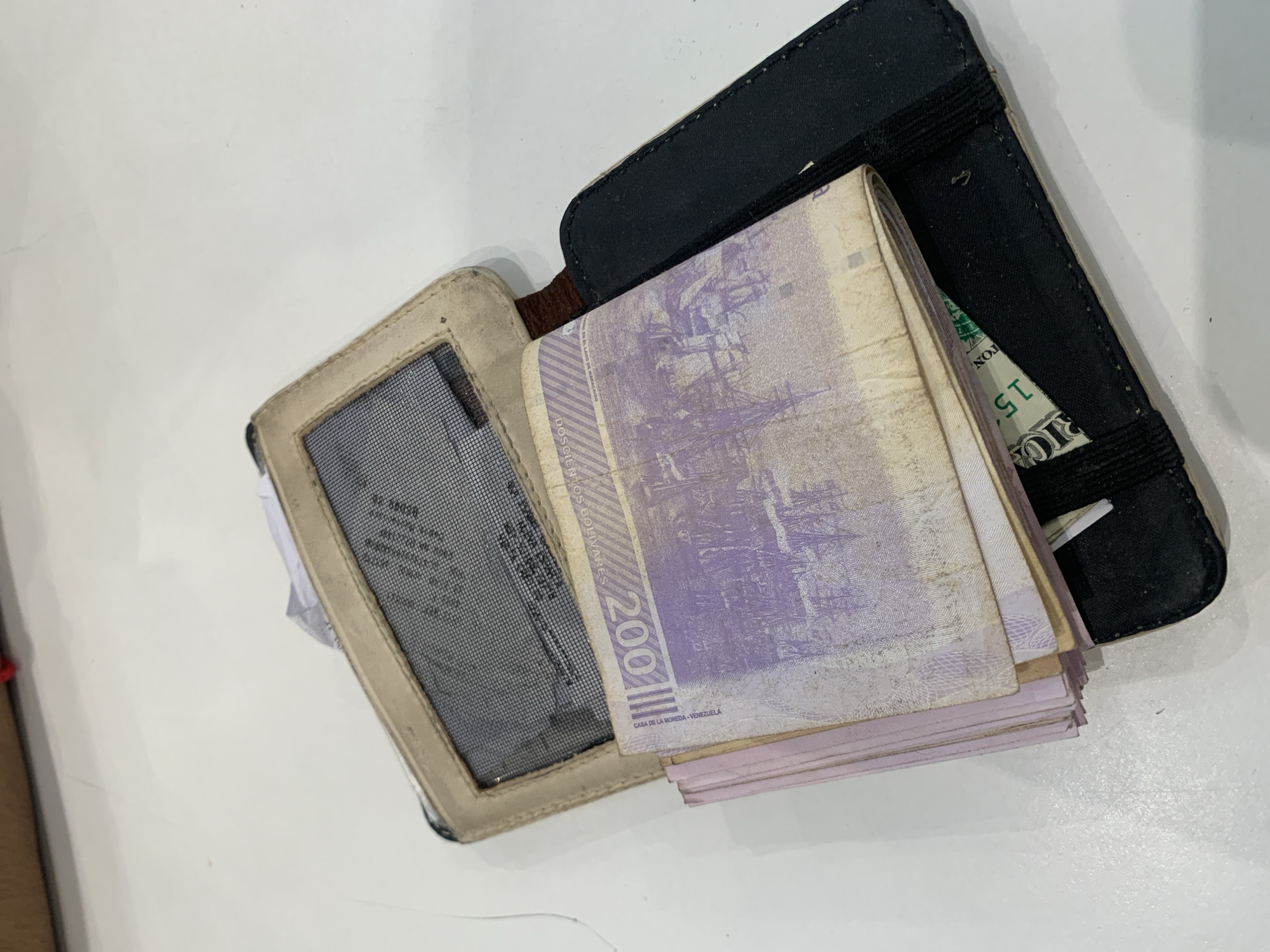 An open wallet, consisting of a black outer section and a beige/tan inner section with a mesh pocket, is presented on a plain, light-colored surface. The wallet is filled with a substantial stack of banknotes, primarily light purple/lavender in color, which appear worn and well-used. A single US dollar bill is also visible, tucked into the black section of the wallet, partially obscured by the stack of local currency.
Notable details include:
- The purple banknotes clearly show the denomination "200" in large numbers, and "DOSCIENTOS BOLIVARES" written vertically. Below the large "200", the text "CASA DE LA MONEDA - VENEZUELA" is visible, indicating these are Venezuelan Bolivars. The imagery on these notes depicts historical structures or ships.
- The US dollar bill shows parts of text like "RIC" (likely from "AMERICA") and "TON", along with the upside-down number "15", which is probably part of its serial number or security features.
- The stack of Venezuelan currency is very thick, suggesting a large quantity of bills.
- The wallet itself shows signs of use, with some scuffs and wear.
- The mesh pocket of the wallet contains some unreadable text on a document or card.
The scene depicts a wallet containing a mix of local Venezuelan currency and US dollars, a common sight in areas with economic instability, consistent with the location context of Pampatar, Venezuela. No specific activity is occurring; it is a static shot of the wallet's contents.