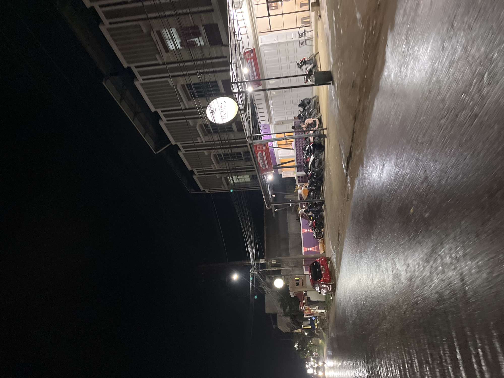 This image shows a street scene at night in Kota Kuala Simpang, Indonesia. A wet road reflects the lights from a row of modern buildings. In front of the buildings, motorcycles are parked. A red car is stopped on the side of the road. The sky is dark, and overhead power lines are visible. There are a few people visible standing near the motorcycles. The overall impression is a quiet, commercial street at night, possibly after rain.