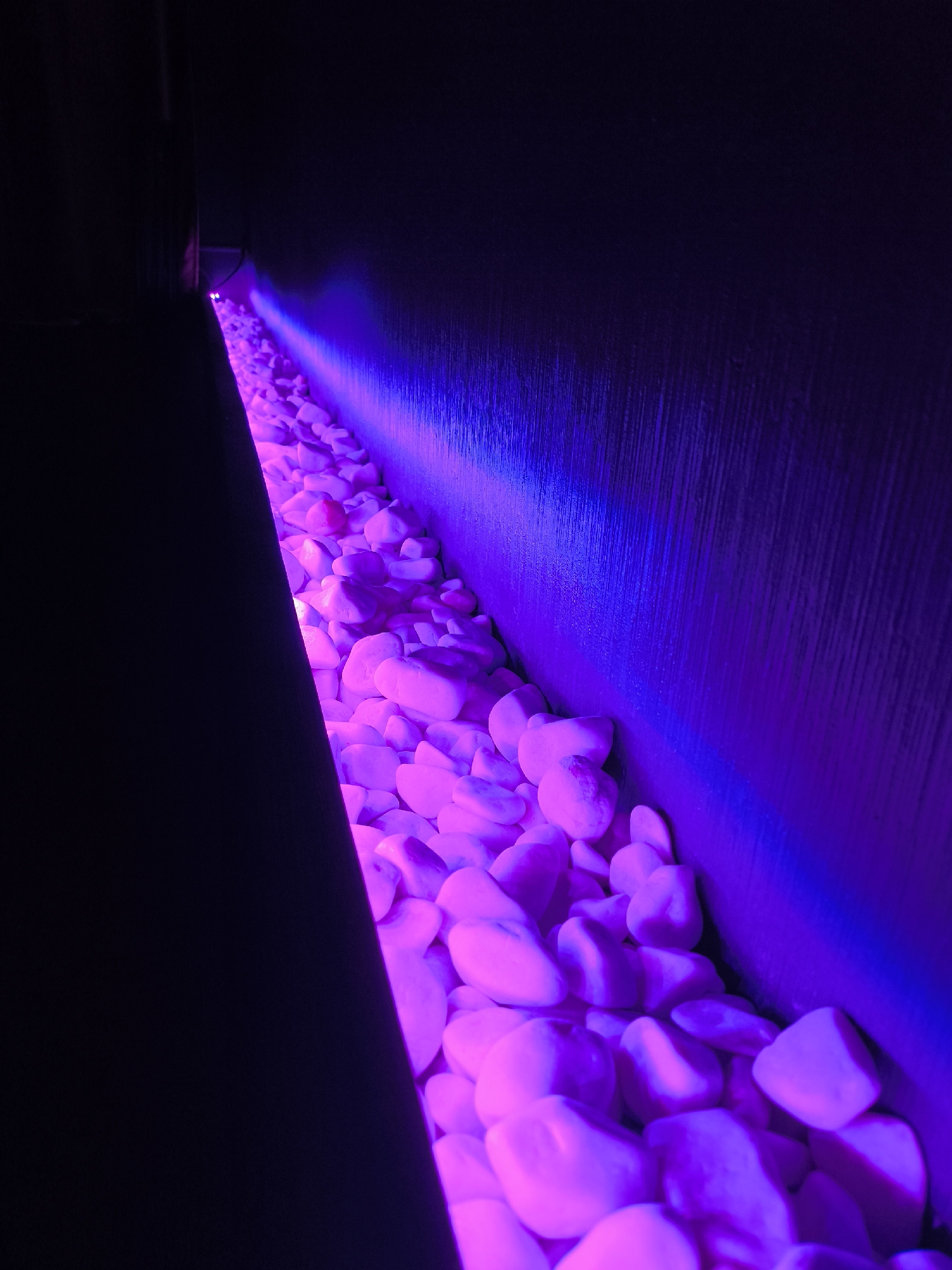 The image shows a close-up of a shallow channel filled with smooth, white stones. The channel is illuminated from above with purple and blue LED lights, casting vibrant reflections on the stones and the dark, textured wall that forms the background. The lighting creates a dramatic, moody effect. The scene is devoid of any people or identifiable urban features, making its specific location in "Unknown, Unknown" impossible to ascertain. The perspective is low, emphasizing the texture of the stones and the gradient of light.