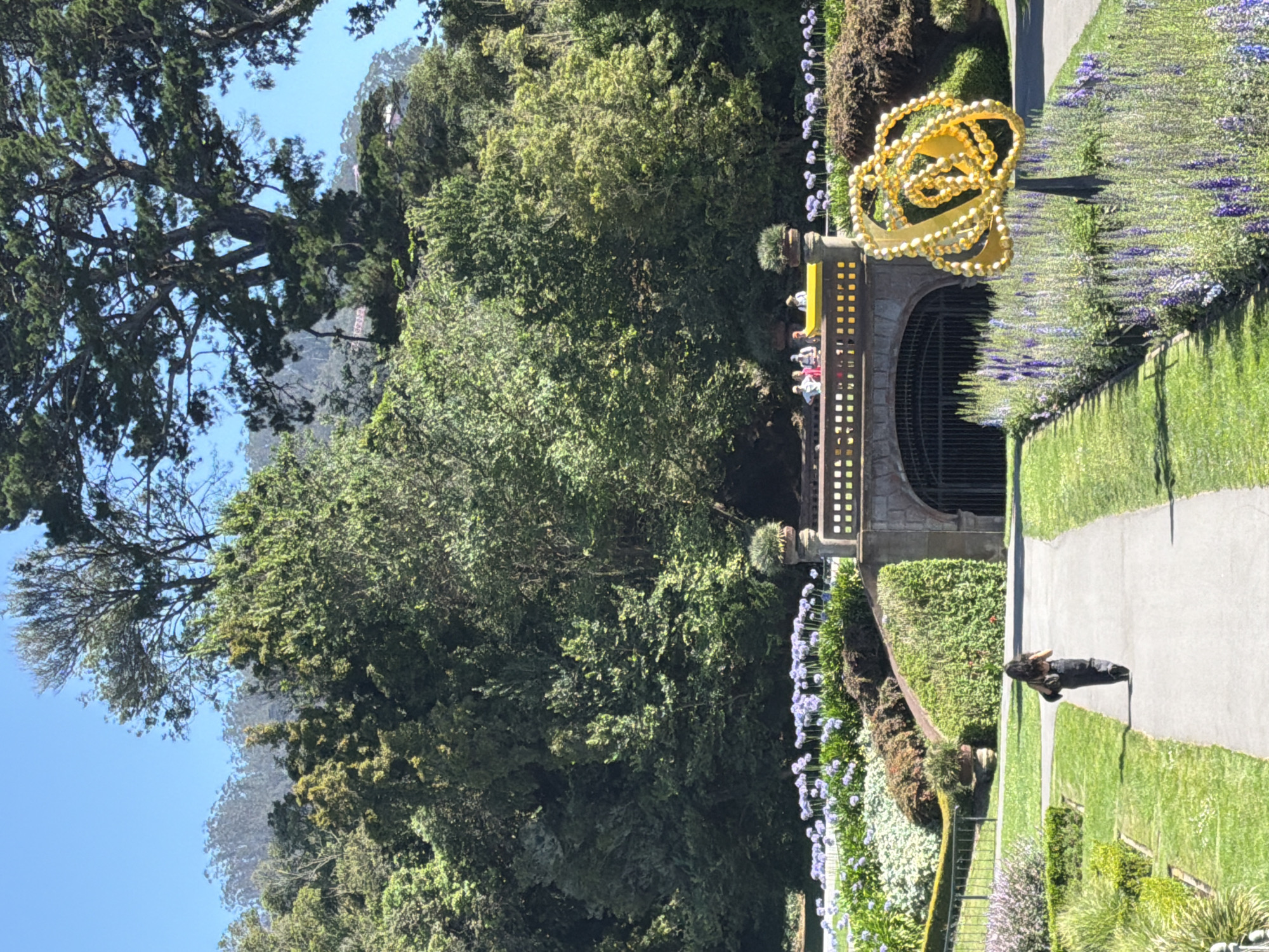 The image depicts a pathway leading towards a stone archway in the Golden Gate Park, San Francisco, California, USA. A lone woman, viewed from behind, walks away from the camera on the pathway. To the right of the pathway, a bright yellow, ornate sculpture stands prominently amidst a bed of purple flowers. In the background, a bridge with people standing on it spans over the archway. Lush green trees and foliage surround the scene, with a clear blue sky above.