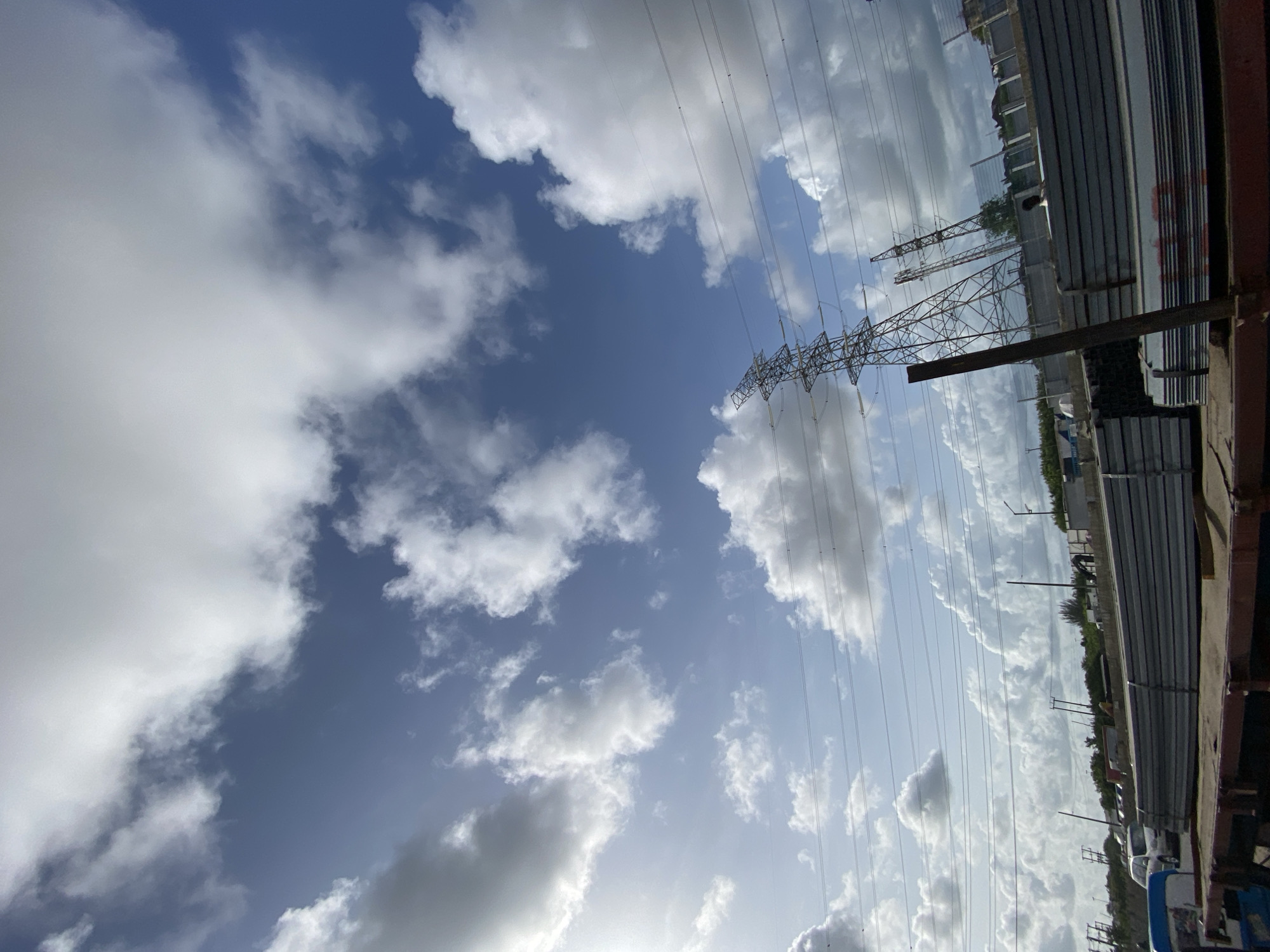 The image shows a view in Lekki, Nigeria, under a sky with scattered white and gray clouds and visible blue sky. Electrical power lines and support structures stretch across the frame, and stacks of long, rectangular metal objects are visible on the right. The image is taken from a relatively low vantage point. The overall impression is of an industrial or urban setting with infrastructure.