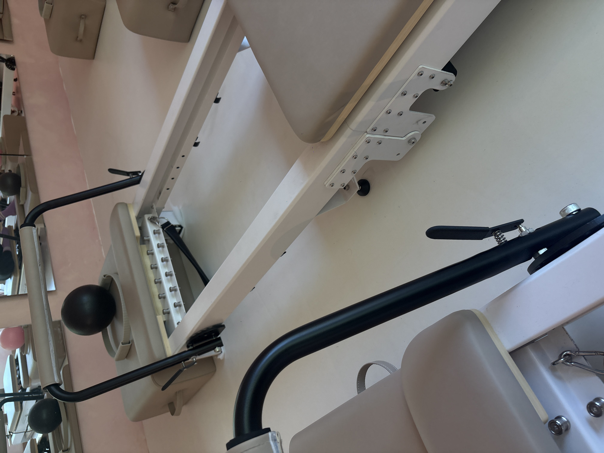 The image shows a partial view of what appears to be physical therapy or exercise equipment. The equipment is constructed of metal and beige-colored padding. A black bar with a lever and spring is visible in the foreground, positioned above a padded section. The background is a light beige, and there is a glimpse of a row of hanging bags and objects, suggesting a shop or store environment, possibly in Mexico City.