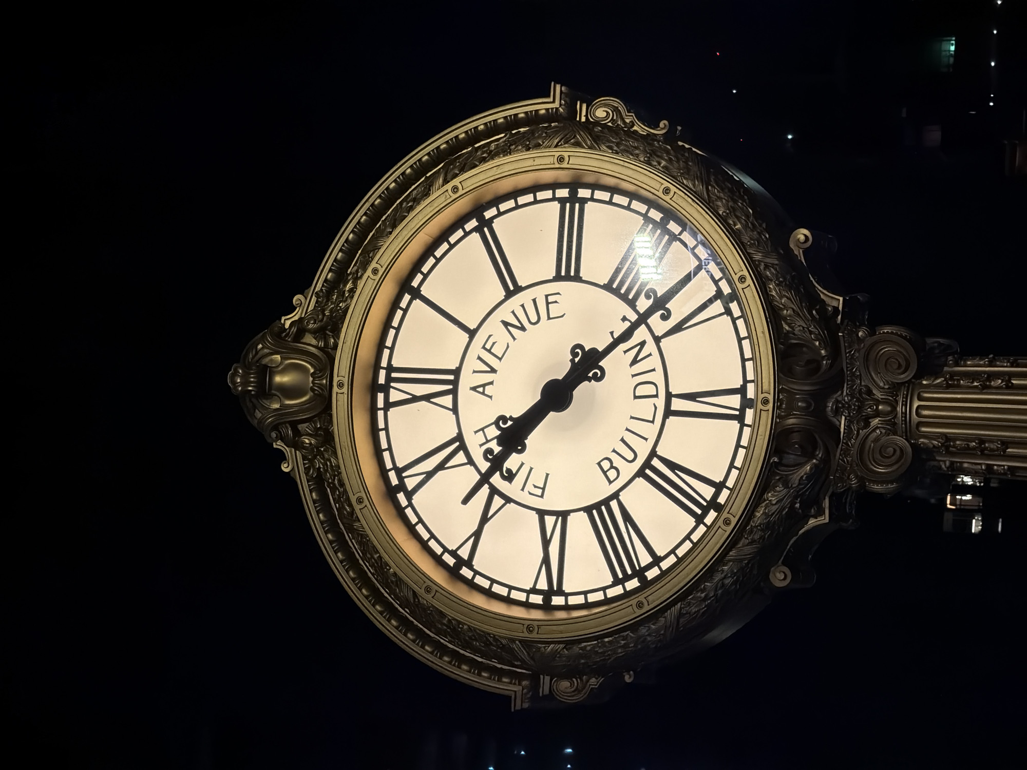 The image is a medium shot of a large, ornate, illuminated clock located on a building. The clock face is white with Roman numerals and has the words "FIFTH AVENUE BUILDING" written around the center. The clock has two hands, with the hour hand pointing to approximately 10:53 and the minute hand pointing to approximately 11:00. The clock is framed by a decorative gold border and is mounted on a dark, ornate pedestal. The background is dark and appears to be nighttime, with some distant lights visible. The building is likely in New York City, given the "Fifth Avenue" inscription.