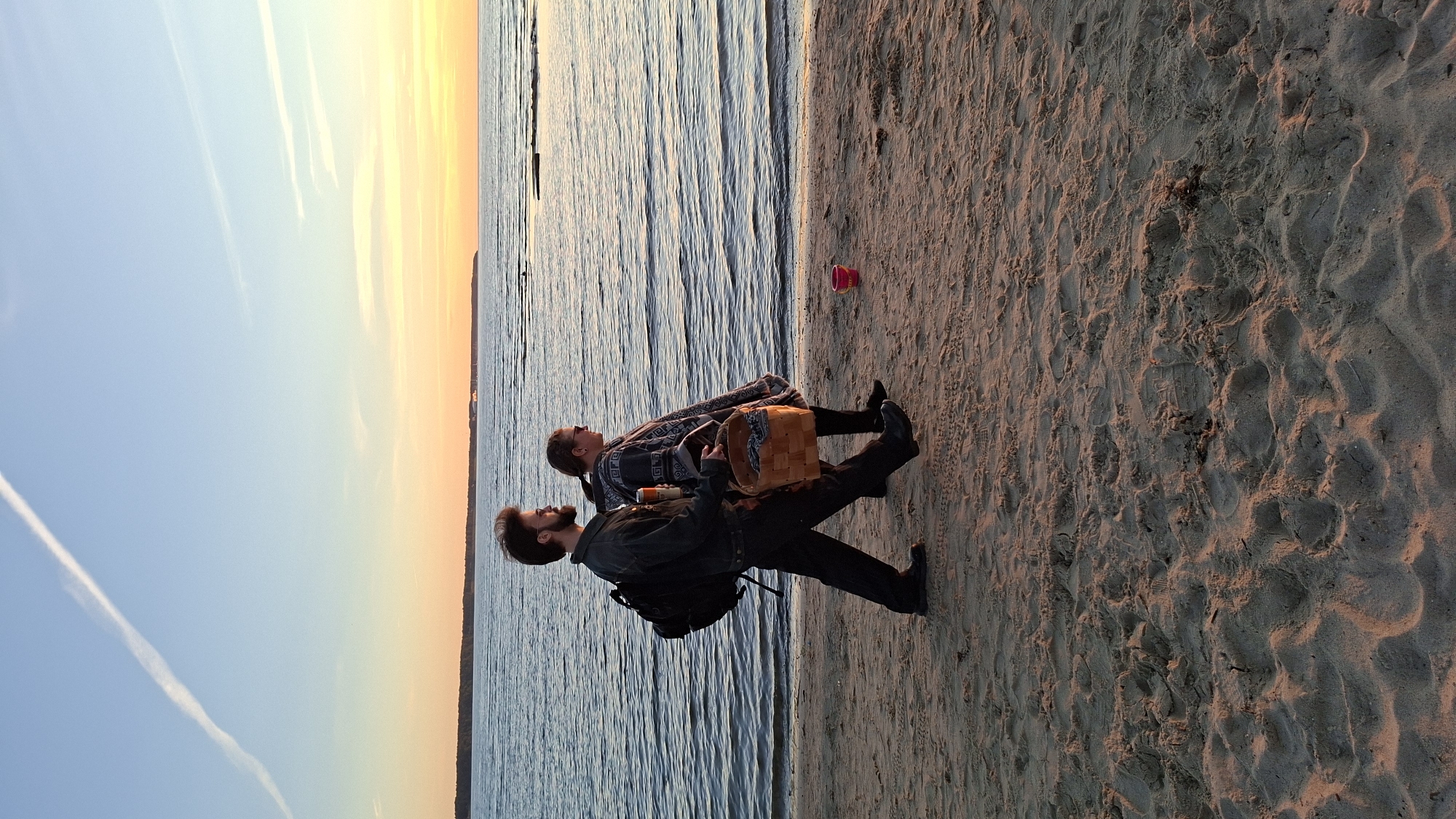 A man and a woman are walking on a sandy beach next to calm water during sunset. The man, on the left, is wearing a dark jacket, dark pants, and black shoes. He has a backpack on his back and is carrying a can in his right hand. The woman, on the right, is wearing a patterned poncho and black pants. She is carrying a woven basket in her arms, which appears to contain a patterned cloth. A small pink bucket is visible on the sand to the right of the couple. The sky is clear with soft hues of orange and yellow near the horizon, and a thin white contrail stretches across the blue sky. The water is a deep blue, reflecting the colors of the sunset. The sand is light brown and textured with footprints and small mounds. The scene appears to be a public beach. The location is not specified in the image, but if this were Tallinn, Estonia, it could be a beach like Pirita or Kakumäe.