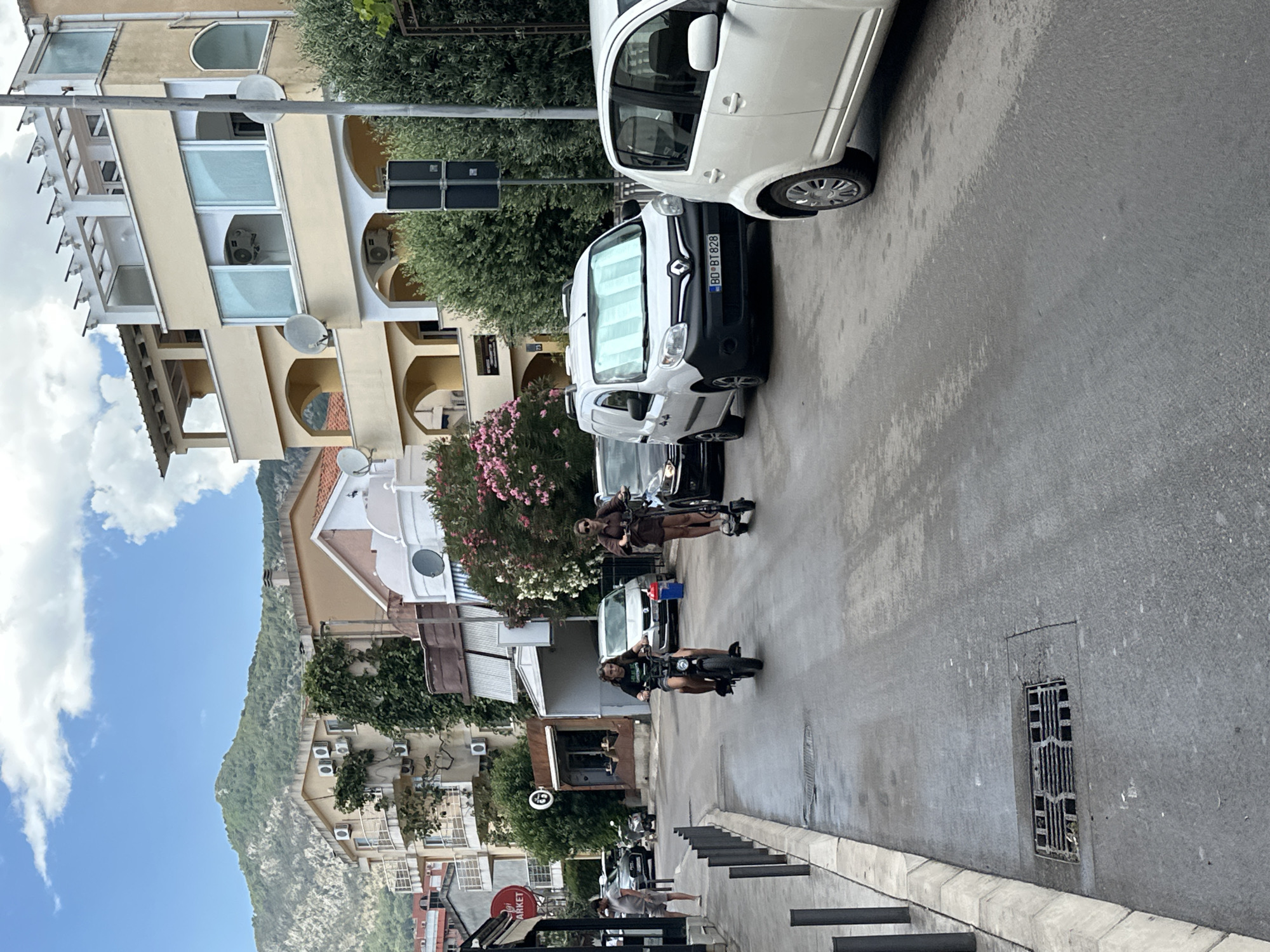 The image depicts a street scene in Budva, Montenegro. In the foreground, a gray asphalt road is wet, suggesting recent rain. A storm drain is visible on the right side of the road. Two individuals are riding personal electric vehicles on the road; one is on a scooter-like device, and the other is on a more substantial electric scooter or moped. Both are dressed casually.
Several vehicles are present, including a white van with a license plate starting with "BD BT 828", a white car, and a dark-colored car. In the background, buildings with multiple stories line the street. Some buildings have balconies and air conditioning units. Green foliage and trees are interspersed with the architecture. Further in the distance, a lush green mountain dominates the skyline under a partly cloudy sky. The overall impression is of a moderately busy street in a coastal town with a mountainous backdrop.