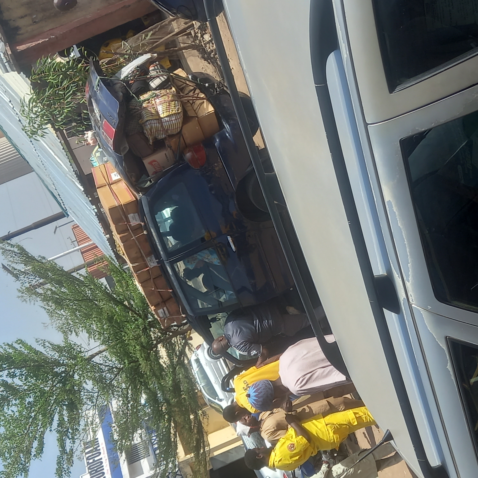 The image depicts a bustling outdoor scene in Dadi Riba, Nigeria, under bright, clear daytime skies. A dark blue utility vehicle, possibly a pick-up truck or SUV, is prominently featured, heavily loaded with goods. Its roof and rear cargo area are piled high with numerous cardboard boxes, some clearly labeled "Milla," along with several striped fabric bundles and green leafy branches.
Around the vehicles, several men are present. One man, dressed in a dark grey or black shirt, is seen leaning into the interior of the dark vehicle. Near a lighter-colored car, partially visible to the right, a group of men are gathered. Two men wear bright yellow shirts, one with a blue patterned cap, and another wears a light pink shirt over a brown garment. Their postures suggest they are actively involved in loading, unloading, or observing the process.
In the background, lush green trees are visible, and behind them, a building with a blue and white sign can be partially seen. The sign displays the prominent text "UROFOAM," with smaller, partial words like "TO" and "JOGAO" also discernible. The overall scene conveys a busy, commercial atmosphere.