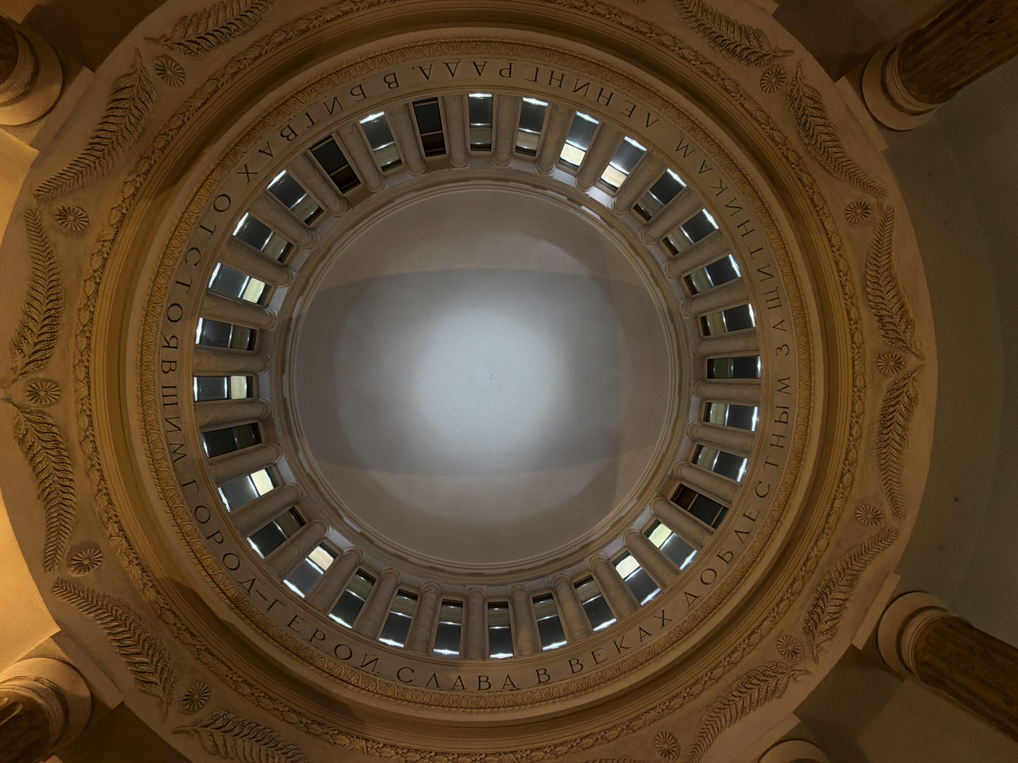 The image shows the interior of a dome in Saint Petersburg, Russia. A ring of window-like openings encircles the dome's base, and between them are evenly spaced, vertical support beams. The inscription above the windows is in Russian. The surface of the dome is uniformly pale. The architecture features ornate detailing, including leaf and flower-like patterns. Columns are visible at the periphery of the image, indicating the building's larger architectural structure.
