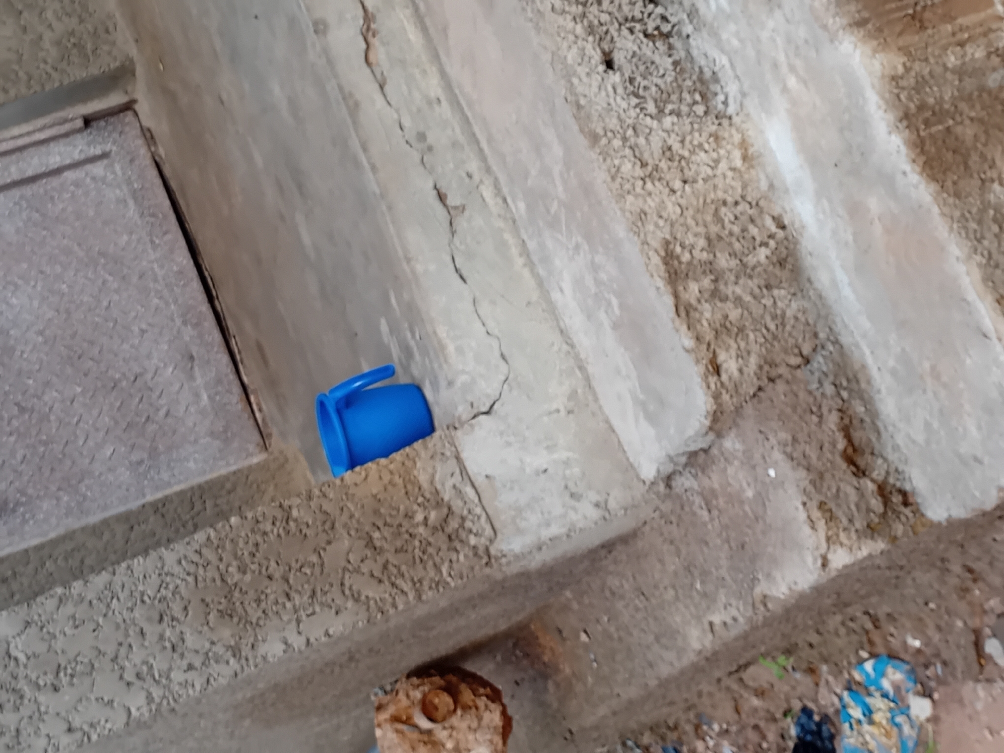 A blue plastic mug sits on a cracked concrete step in Jauro Yinu, Nigeria. The mug is positioned in the foreground to the right, with its handle facing right. The step is part of a set of tiered concrete steps leading up towards a weathered metal door visible in the upper right portion of the image. To the left of the steps, the textured concrete wall of a building is visible, with some debris on the ground below it. The overall scene appears to be outdoors, possibly in an alley or near an entranceway.