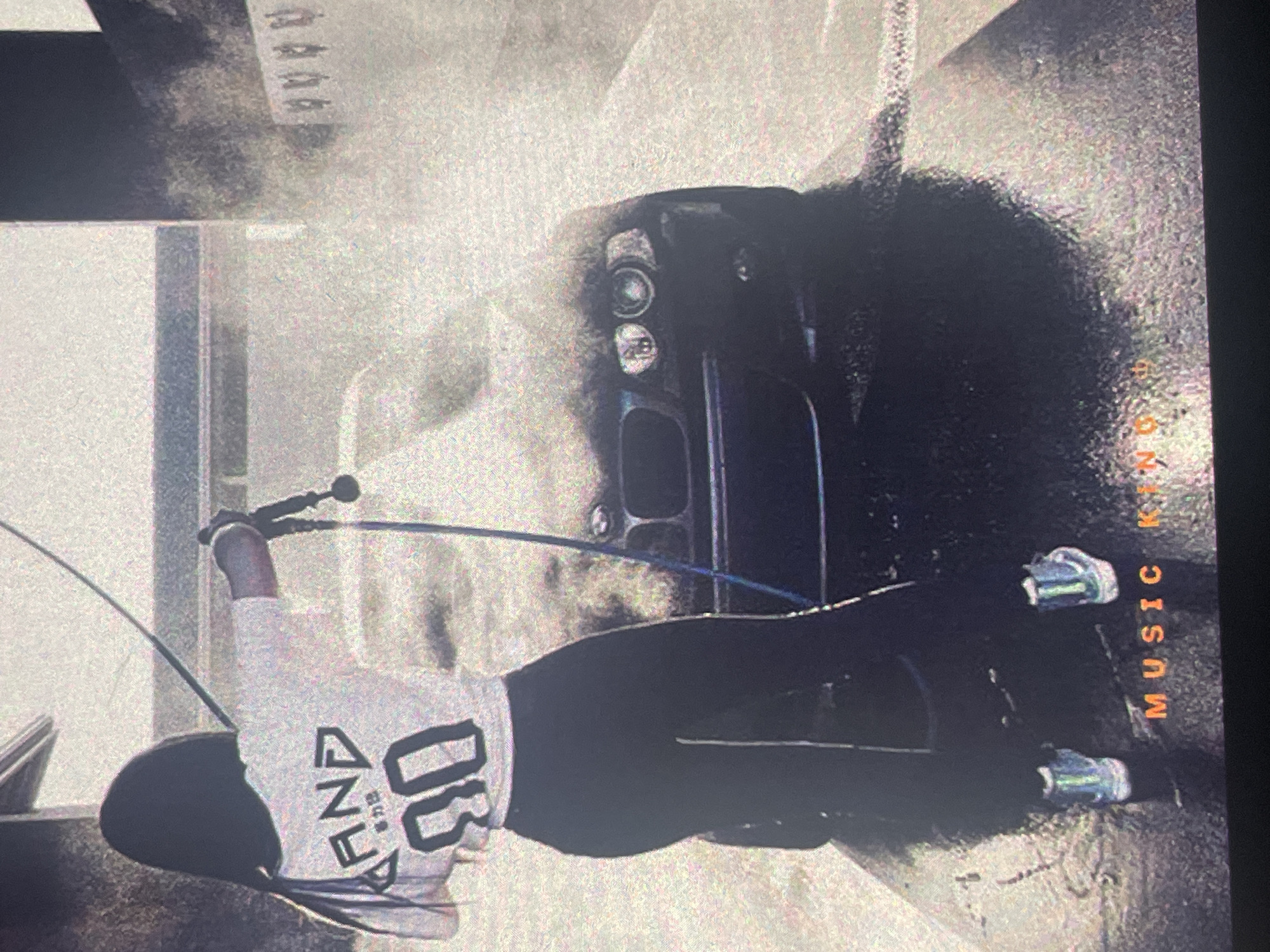 The image depicts a woman washing a black car at a car wash. She is wearing a white t-shirt with Japanese characters and the number "90" on the back, and black leggings. She is holding a high-pressure washer and spraying water on the car. The car wash appears to be an outdoor facility with a concrete floor. The background shows a building structure and some white railings. The image also has text at the bottom that reads "MUSIC KINGDOM". The scene is likely set in Paczkowo, Poland, given the context.