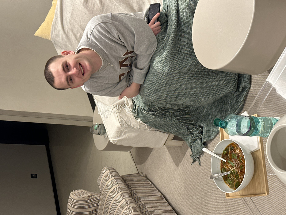 A man sits on a couch covered in a patterned green blanket, holding a phone. He's wearing a gray sweatshirt. A white side table holds a bowl of soup with two spoons in it, a bottle of water, and a white pitcher. The couch is off-white with a yellow pillow. Another striped couch is partially visible in the foreground. A door is visible on the left.
