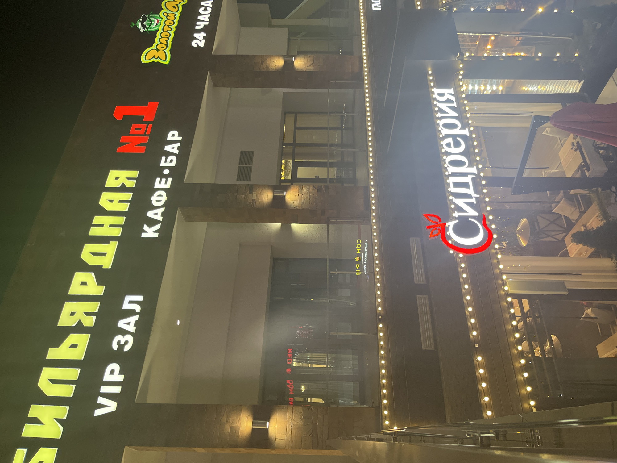 The image depicts the exteriors of two restaurants, located in Sochi, Russia. On the left is a restaurant named "БИЛЬЯРДНАЯ No. 1", with the words "КАФЕ-БАР" and "VIP ЗАЛ" underneath. Above this is a logo featuring the words "ЗОЛОТОЙ ОСЕЛ" along with the text "24 ЧАСА". To the right is another restaurant called "Сидрерия". Strings of lights adorn the exteriors of both establishments. The restaurants appear to be located in a modern building complex with several stories and external architectural details like brickwork and recessed lighting. Inside "Сидрерия," tables and chairs are visible, suggesting a seating area. The overall scene is captured at night, with the artificial lights illuminating the establishment.
