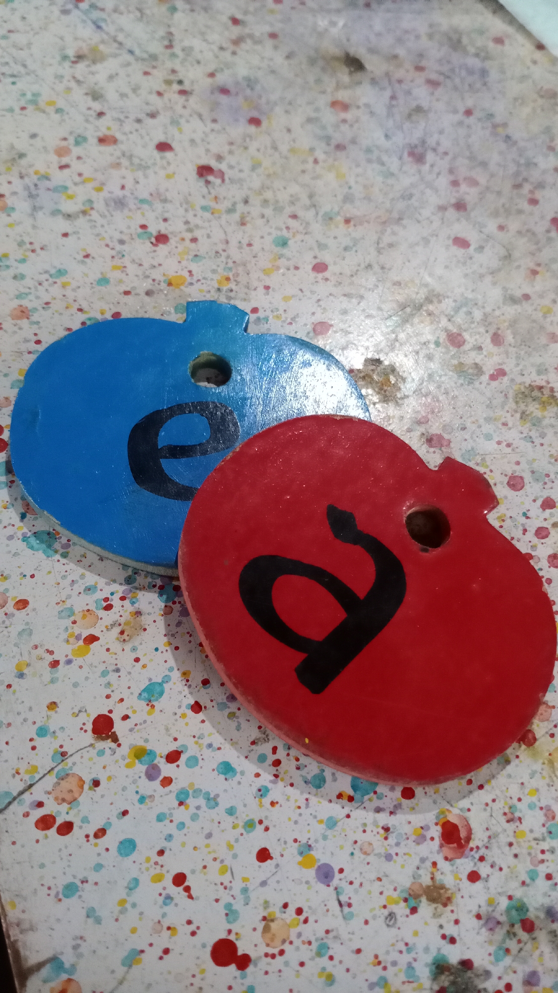 Two apple-shaped wooden ornaments are displayed on a speckled surface. The ornament in the foreground is red and features a black letter "a." The ornament behind it is blue and has a black letter "e." Both ornaments have a small hole at the top, suggesting they might be used as tags or decorations. The background surface is white with colorful paint splatters in red, yellow, blue, and purple. There are no people or animals visible in the image. The image does not provide any information about the location in the city of Mertan, Indonesia.