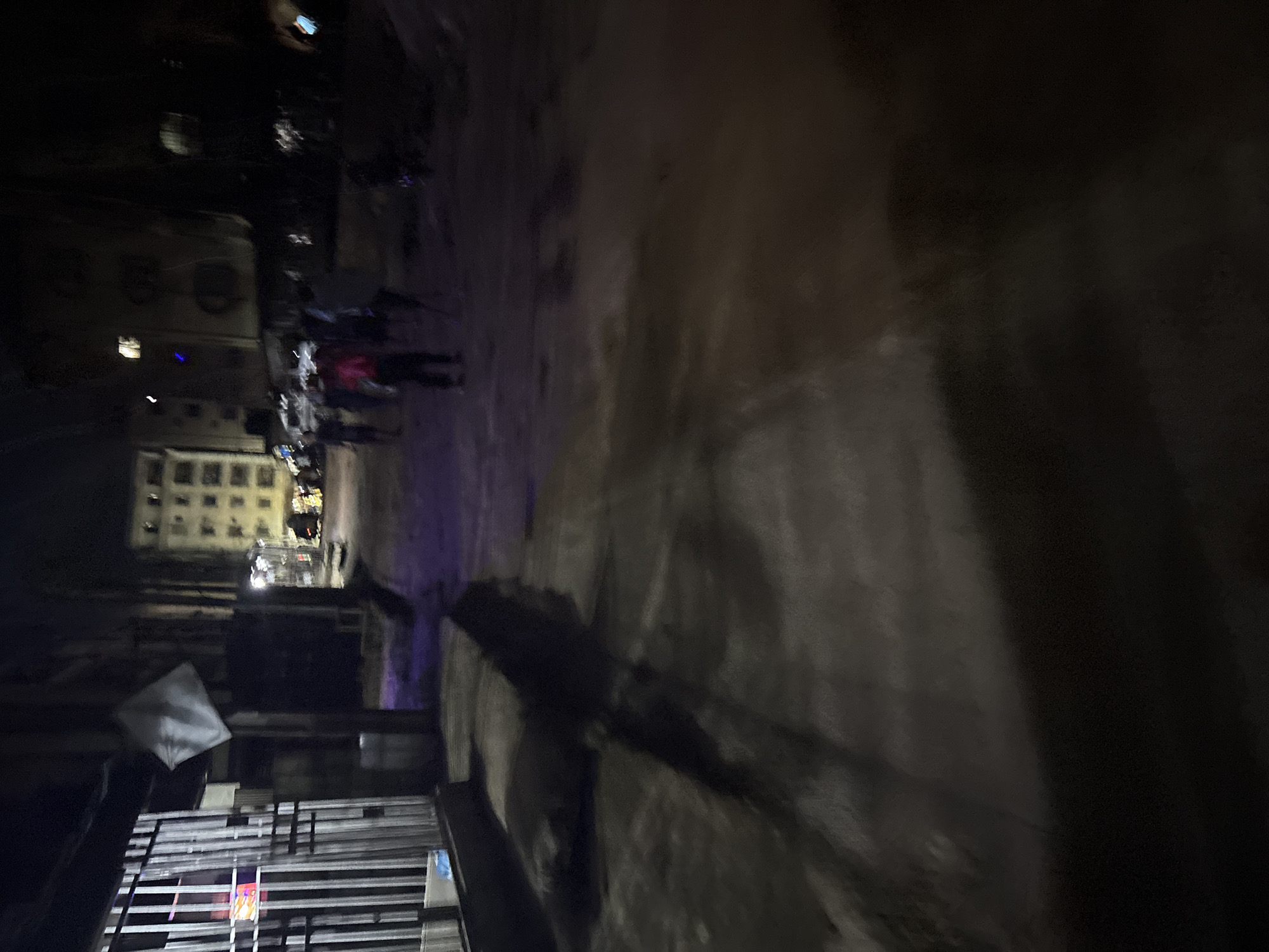 The image depicts a nighttime street scene in Onitsha, Nigeria. A metal gate with vertical bars is visible on the left side of the frame, with some colorful lights or advertisements visible behind it. A white, diamond-shaped object is mounted on a pole in front of the gate. The street appears to be unpaved and uneven, with shadows cast on the ground.
In the background, several buildings are dimly lit. One building is a multi-story structure with many windows, appearing to be an apartment complex or hotel. Other structures are also visible, but less distinct due to the low light conditions.
A group of people are walking or standing in the street. One person in the foreground is wearing a pink top and dark pants. Other individuals are less clearly defined, appearing as silhouettes against the background lights. Some are walking away from the camera, while others are facing towards it. The overall atmosphere suggests a busy urban environment at night, with limited illumination. The presence of the gate and the unpaved street indicate a location within a residential or commercial area, possibly with informal settlements.