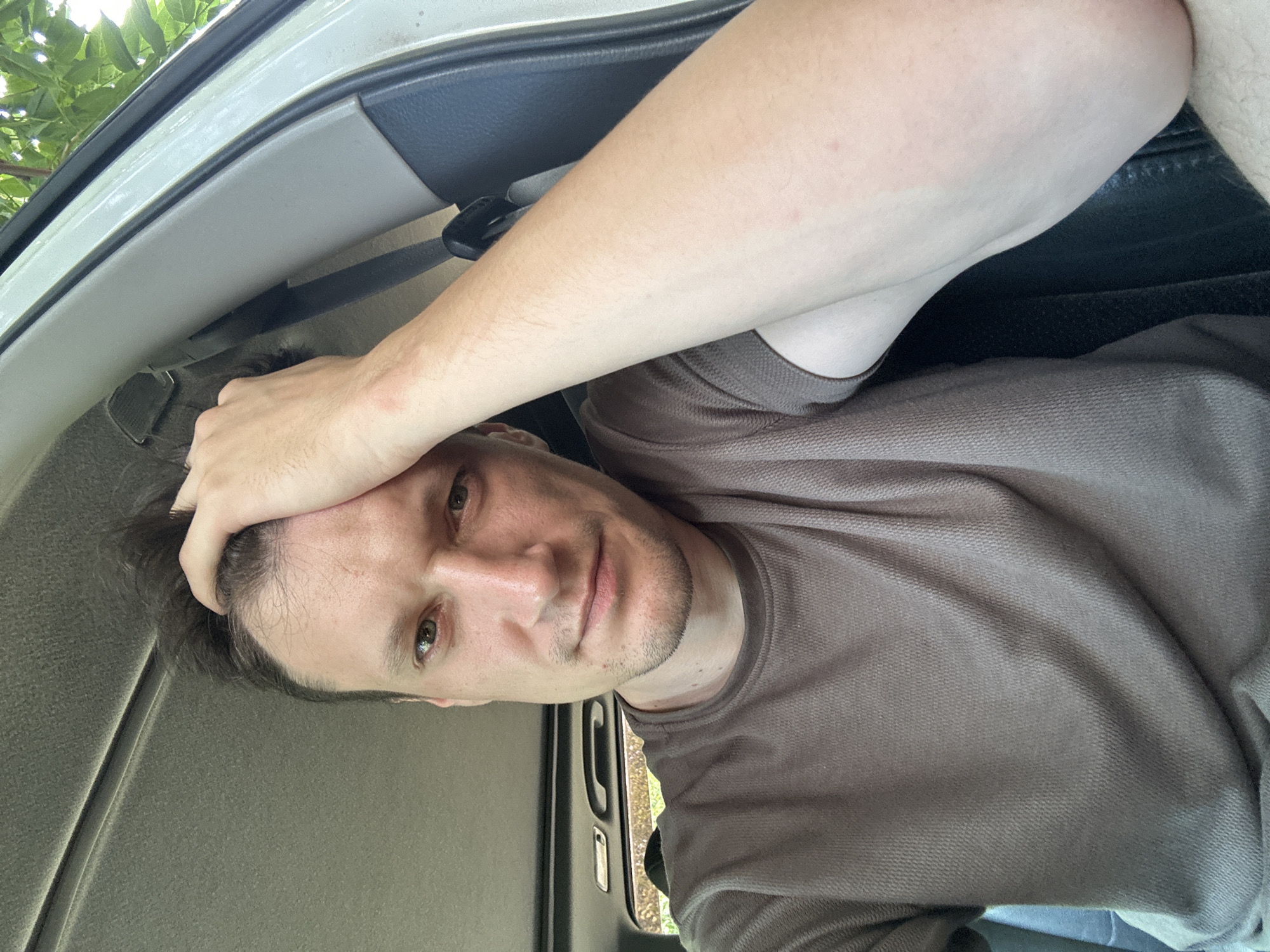 In a selfie taken from inside a vehicle, a man with light skin and short, dark brown hair is seen with his right hand on his head. He is wearing a dark grey t-shirt. The man is positioned in the driver's seat of the car, with the interior visible, including the car's headliner and a portion of the dashboard. Outside the car, greenery and trees are visible, suggesting a natural or outdoor setting. The lighting indicates it is daytime. The exact location in Makiv, Ukraine, is not discernible from the image.