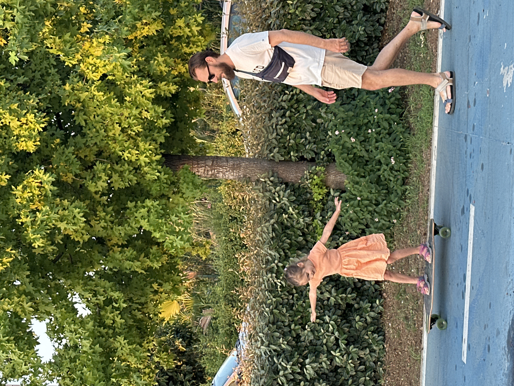 A young girl is skateboarding on a blue-surfaced court, her arms outstretched for balance. She is wearing a peach-colored dress and pink sandals. Behind her, a man with a beard and sunglasses is walking, also with his arms outstretched, wearing a white t-shirt, beige shorts, and dark sandals. A dark pouch is slung across his chest. The setting appears to be outdoors, with lush green trees and bushes framing the scene. The sunlight suggests it is daytime. In the background, there is a glimpse of a white vehicle. The overall mood seems relaxed and recreational. The image also includes a tree trunk. The location is Fethiye, Turkey.
