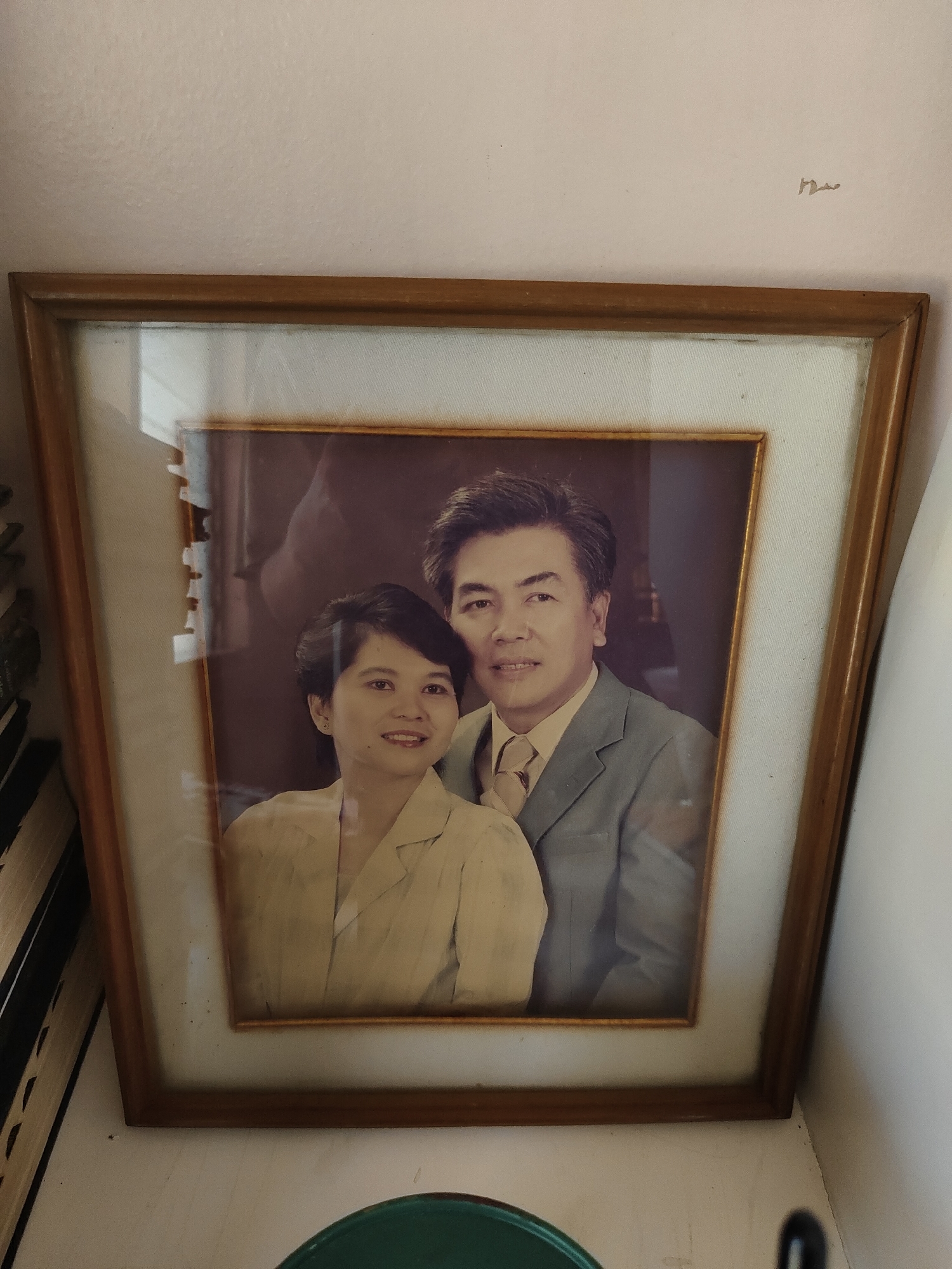 The image displays a framed portrait of a man and a woman, likely a couple, set against a light-colored, textured wall in an indoor setting. The wooden frame encloses a light, off-white matting around the photograph. Both individuals in the portrait appear to be of Southeast Asian descent. The woman, positioned slightly in front and to the left, has short dark hair and a gentle smile, wearing a light-colored, possibly cream or beige, collared blouse. The man, standing behind and to the right, also smiles gently and has dark hair; he is dressed in a light blue or grey suit jacket with a light-colored collared shirt and tie. The background within the portrait is a plain, dark hue.
Reflections from the room are visible on the glass covering the photograph, including hints of a vertical white surface on the left and various light sources. The framed photo rests on a surface. To the bottom left of the frame, several book spines are partially visible, suggesting a shelf or table location. To the bottom right, a segment of a dark green, circular object, possibly a bowl, is seen. The wall above the frame shows some faint marks and texture. The photograph itself has a somewhat vintage aesthetic. There is no visible text in the image. The scene conveys a personal and cherished display, consistent with a home in a location like Davao City, Philippines.