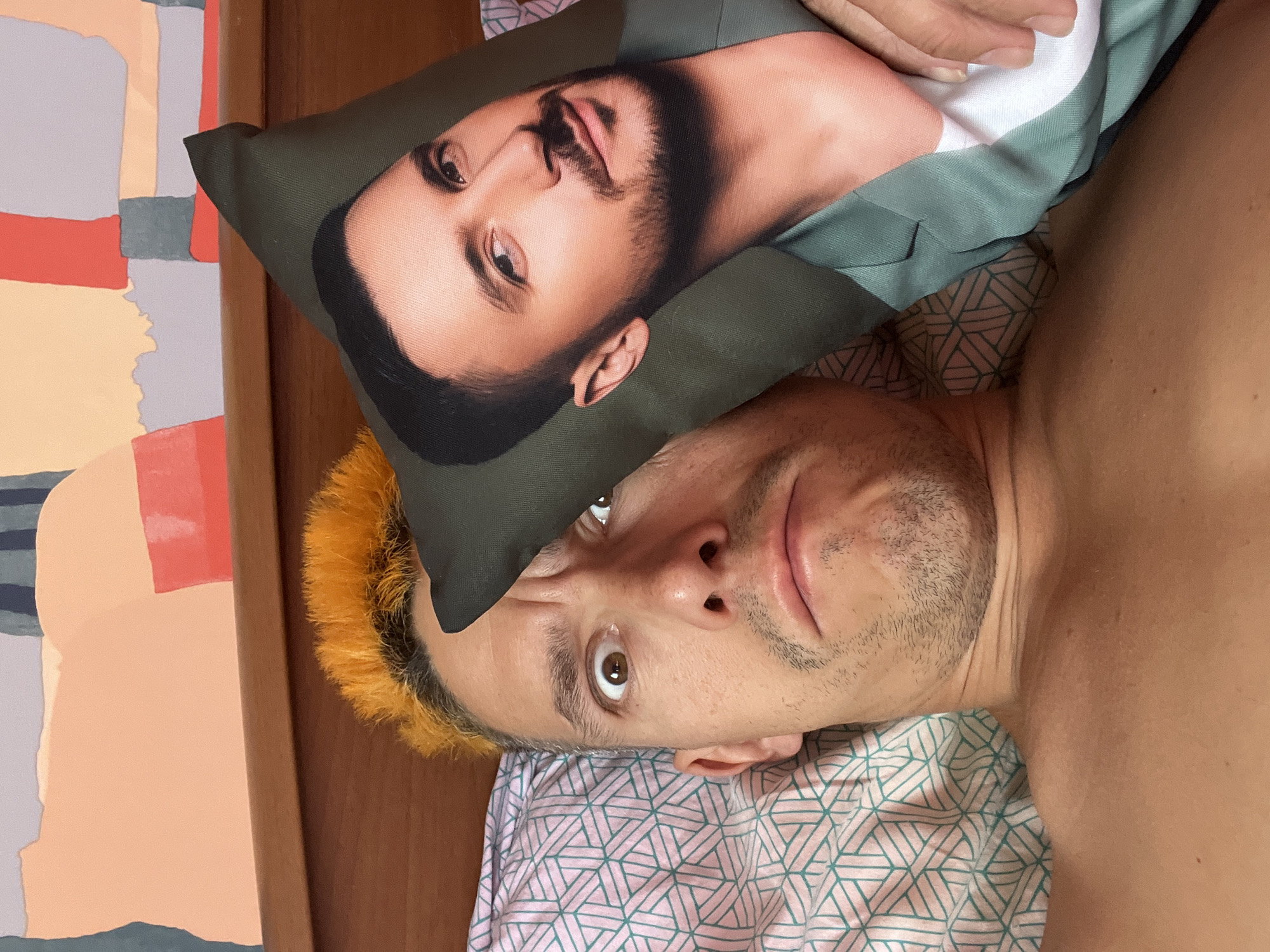 The media file shows a close-up, overhead shot of a person lying in bed. The person has short, brightly dyed orange hair and is looking directly at the camera with wide, surprised eyes. A pillow with a printed image of another person's face is placed on top of the person's head. The pillow has a dark green background and the printed face has stubble and is wearing a green jacket. The person in the foreground is lying on a bed with pink and green patterned sheets. To the left of the bed is a wooden headboard and a wall with abstract art. The lighting suggests it is daytime, with natural light illuminating the scene. The overall mood is humorous and playful, due to the person's expression and the unexpected placement of the pillow. No visible text is present in the image. The location context of Chaikovsky, Russia, is provided.