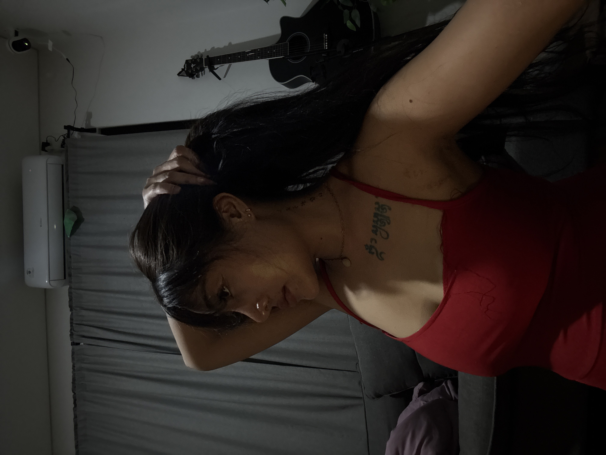 The image captures a person, likely female, with long dark hair, wearing a red sleeveless top, in an indoor setting with dim lighting, suggesting evening or night. The person is seen in a profile view, looking downwards and to the left, with both hands raised to the back of their head as if adjusting or gathering their hair. They have a nose piercing on their left nostril and wear a delicate gold chain necklace with a small, light-colored pendant.
A prominent tattoo with characters resembling an East Asian or Southeast Asian script is visible on their right shoulder/neck area. A smaller, lighter tattoo can be seen on the left side of their neck/chest. A black mole is also visible on their right upper arm.
The background includes a black acoustic guitar hanging horizontally on a light-colored wall in the upper right, adorned with some green foliage. To the left, an air conditioning unit is mounted on the wall, beneath which gray pleated curtains or blinds are visible. A small, black cylindrical device, possibly a projector or camera, is mounted on the wall in the upper left corner, with cables running from it. The lower right portion of the image shows parts of a dark sofa or bed with a purple blanket or fabric. The overall scene conveys a quiet and somewhat pensive mood.