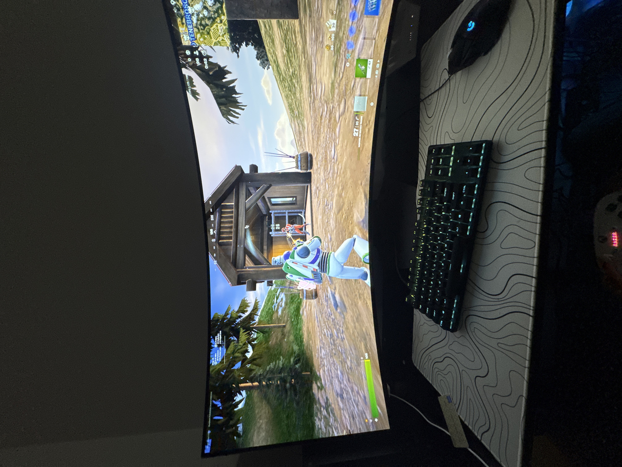 A curved computer monitor displays a Fortnite game with a Buzz Lightyear character running towards a wooden structure. A black keyboard with illuminated keys and a black gaming mouse sit on a mousepad featuring a white topographical map pattern. The monitor, keyboard, and mouse are situated on a dark surface. The location is Glendale Heights, United States of America.