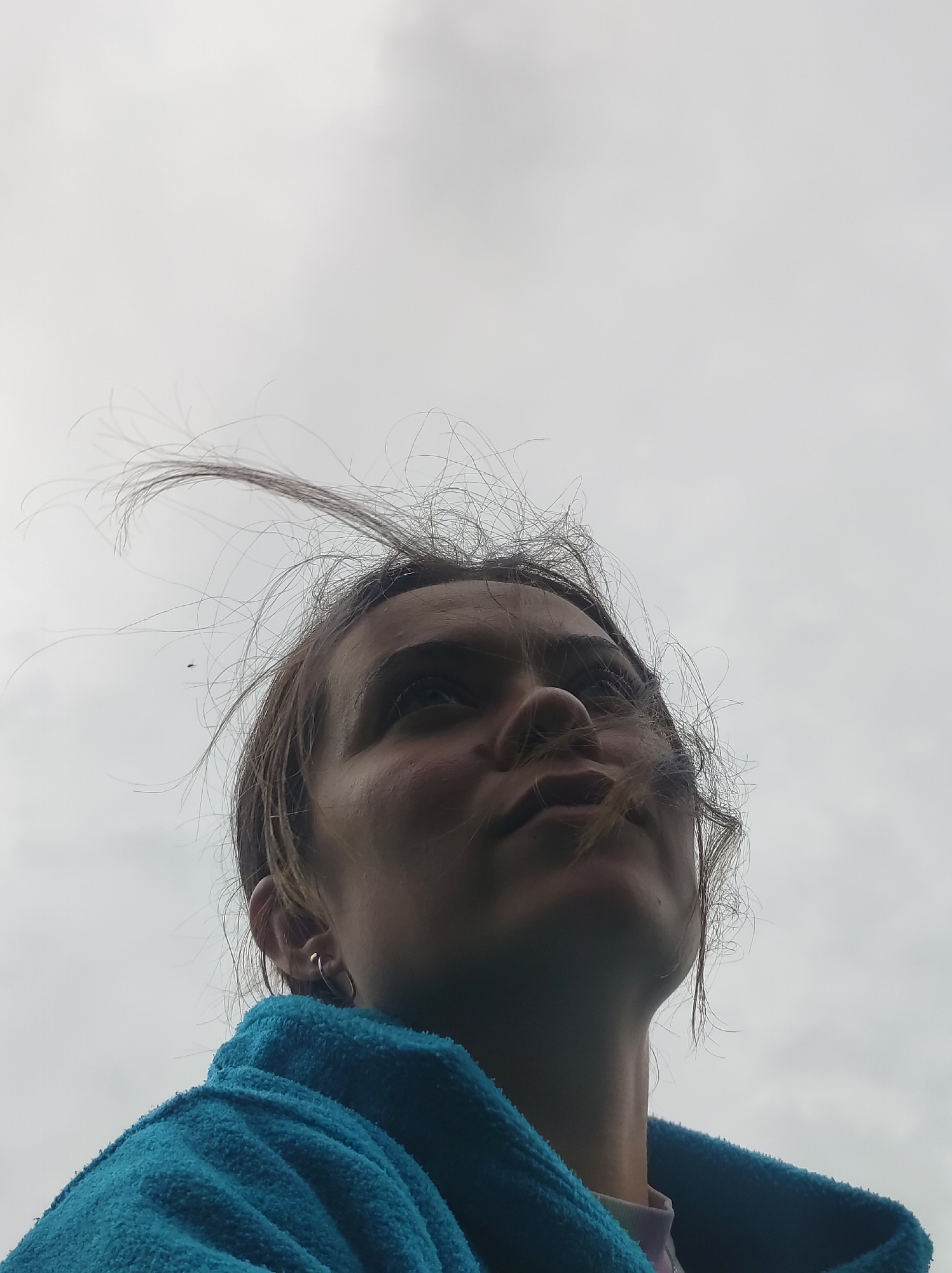 The image shows a close-up, low-angle shot of a young woman from the shoulders up, looking upwards. Her brown hair is windswept, with several strands flying away from her face. She is wearing a bright blue towel or robe. Her right ear is visible with a small hoop earring. The background is an overcast, light gray sky with faint cloud formations. There are no discernible landmarks or elements to suggest a specific location within the city of Фоки, Russia. The lighting is diffused, and the overall mood is somewhat somber or introspective.
