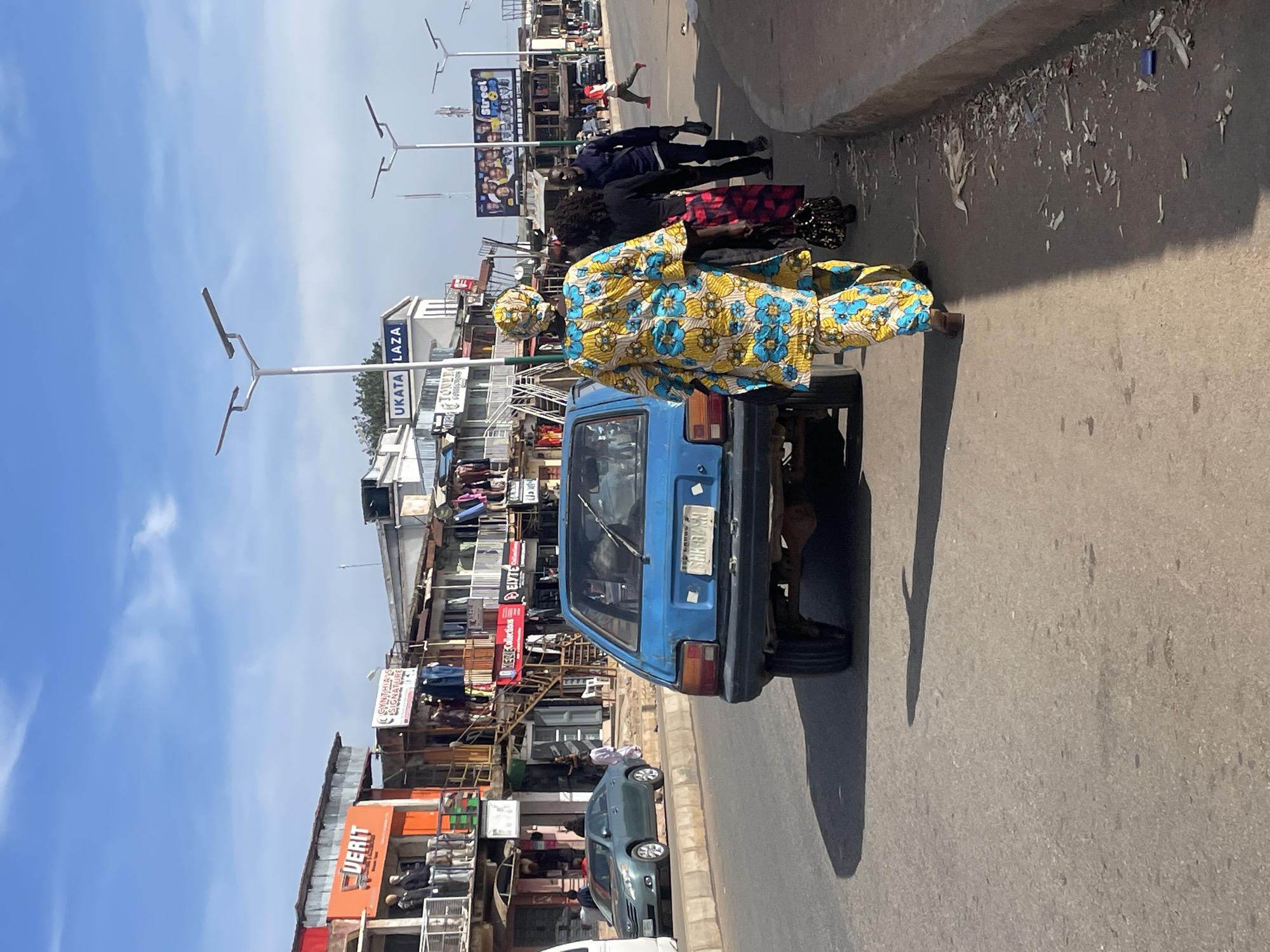 A bright, sunny daytime scene on a paved street in the city of Jos, Nigeria, features a blue hatchback car in the foreground. The car is parked or moving slowly on the right side of the road. Behind the car, three individuals are walking along the sidewalk. The person closest to the car is wearing a vibrant yellow and blue patterned robe and headscarf, walking away from the camera. Next to them, another person is wearing a dark top and a red and black patterned skirt. Further behind, a man in a dark blue shirt and black pants is walking.
On the left side of the street, several multi-story buildings house various businesses. One building has a prominent orange sign that reads "UVERIT" with an image of mannequins on display. Other shops display clothing and merchandise. Several streetlights with solar panels are visible along the street. In the background, a large billboard with images of people and the text "Street" can be seen. The street appears busy with traffic and pedestrian activity, typical of a commercial area in Jos. The ground is paved with asphalt, and the sidewalk is lined with debris. The sky is blue with scattered white clouds.