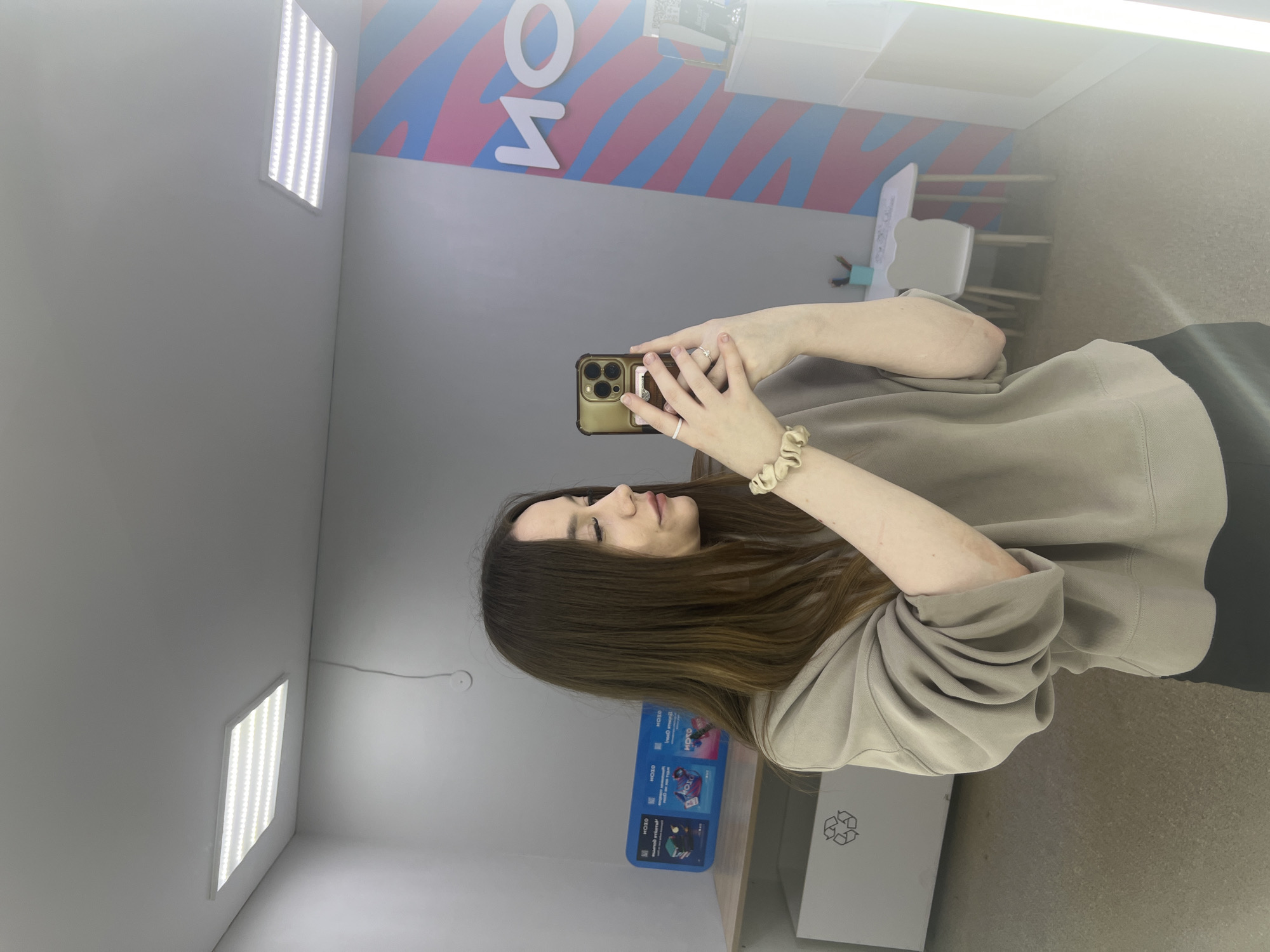 In the city of Chaikovsky, Russia, a young woman with long, brown hair is captured in a mirror selfie. She is wearing a beige sweatshirt and black pants. Her left arm, adorned with a scrunchie, is raised to hold a gold iPhone. The background reveals a room with two rectangular ceiling lights, a shelf displaying blue boxes with white lettering, and a white recycling bin. To the right, a wall features a colorful, wavy design in pink and blue, with white lettering that appears to read "НО". Further to the right, a white table with a child-sized chair is visible. The lighting is bright, with a distinct vertical light source on the far right of the frame.